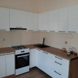 Nowoczesna kuchnia z białymi, lakierowanymi frontami i drewnianym blatem. Widoczny zlew, kuchenka i wiszące szafki z czarnymi, podłużnymi uchwytami. Minimalistyczny design.