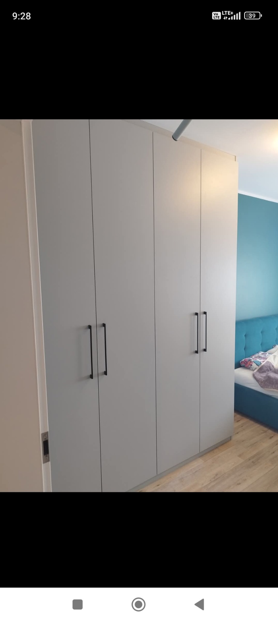 Szara szafa wnękowa z czarnymi uchwytami, widoczna częściowo sypialnia z turkusową ścianą i łóżkiem. Prosty design, jasne wnętrze.