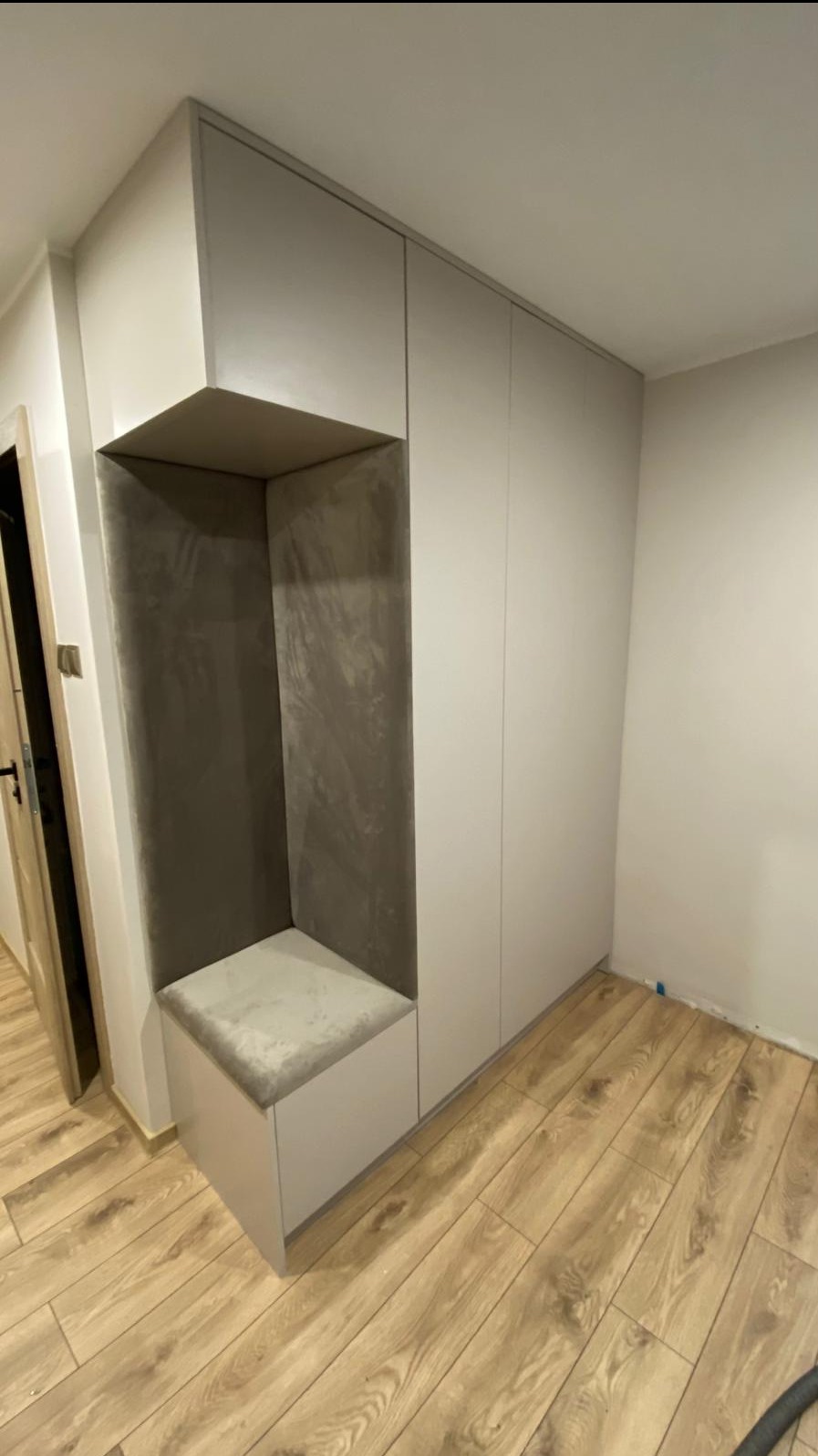 Szara szafa wnękowa z siedziskiem w przedpokoju, drewniana podłoga. Siedzisko z szarą tapicerką. Ściany w jasnym odcieniu szarości. Minimalistyczny design.