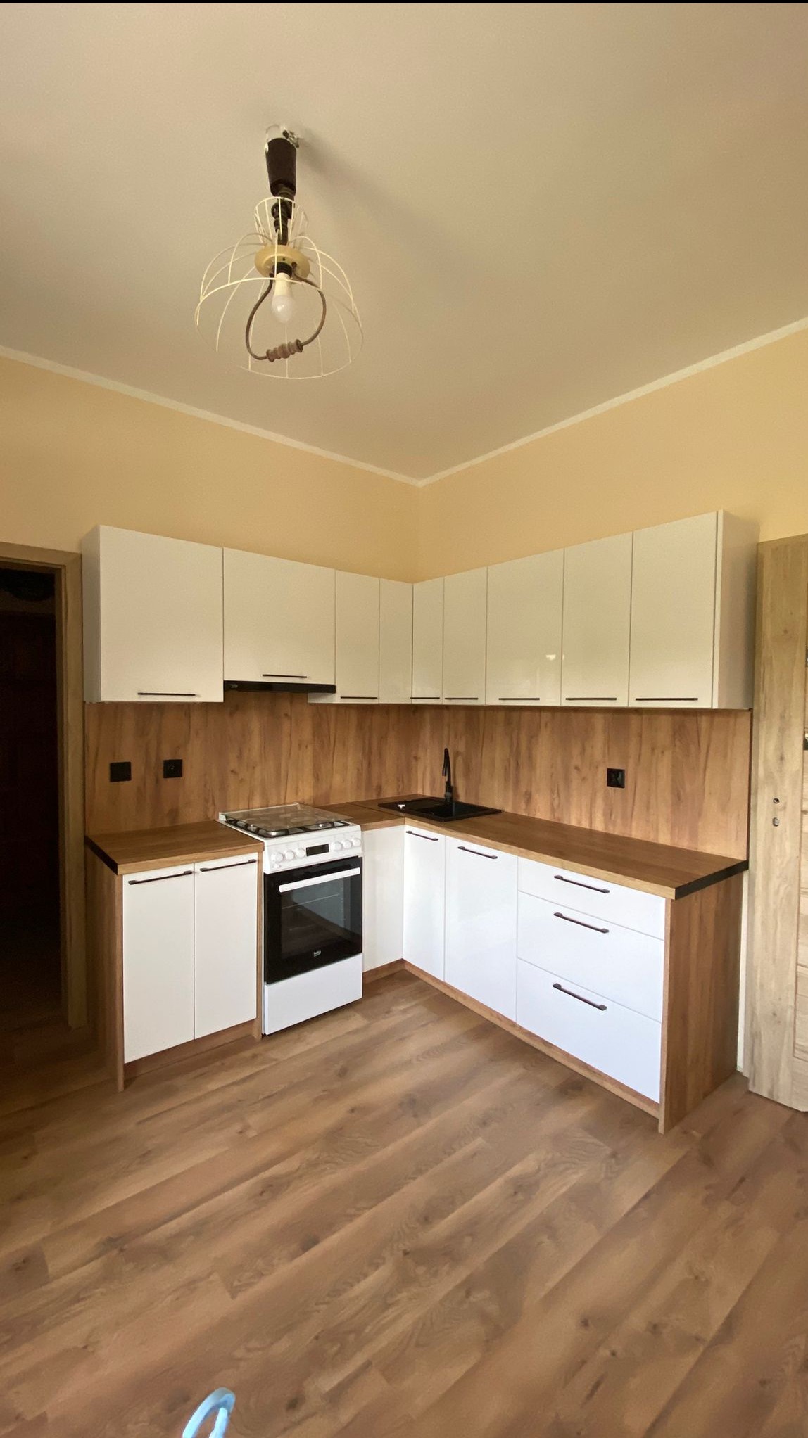 Kompaktowa kuchnia w kształcie litery L: białe szafki z drewnianymi blatami i panelami, czarna armatura, wisząca lampa w stylu industrialnym, drewniana podłoga.