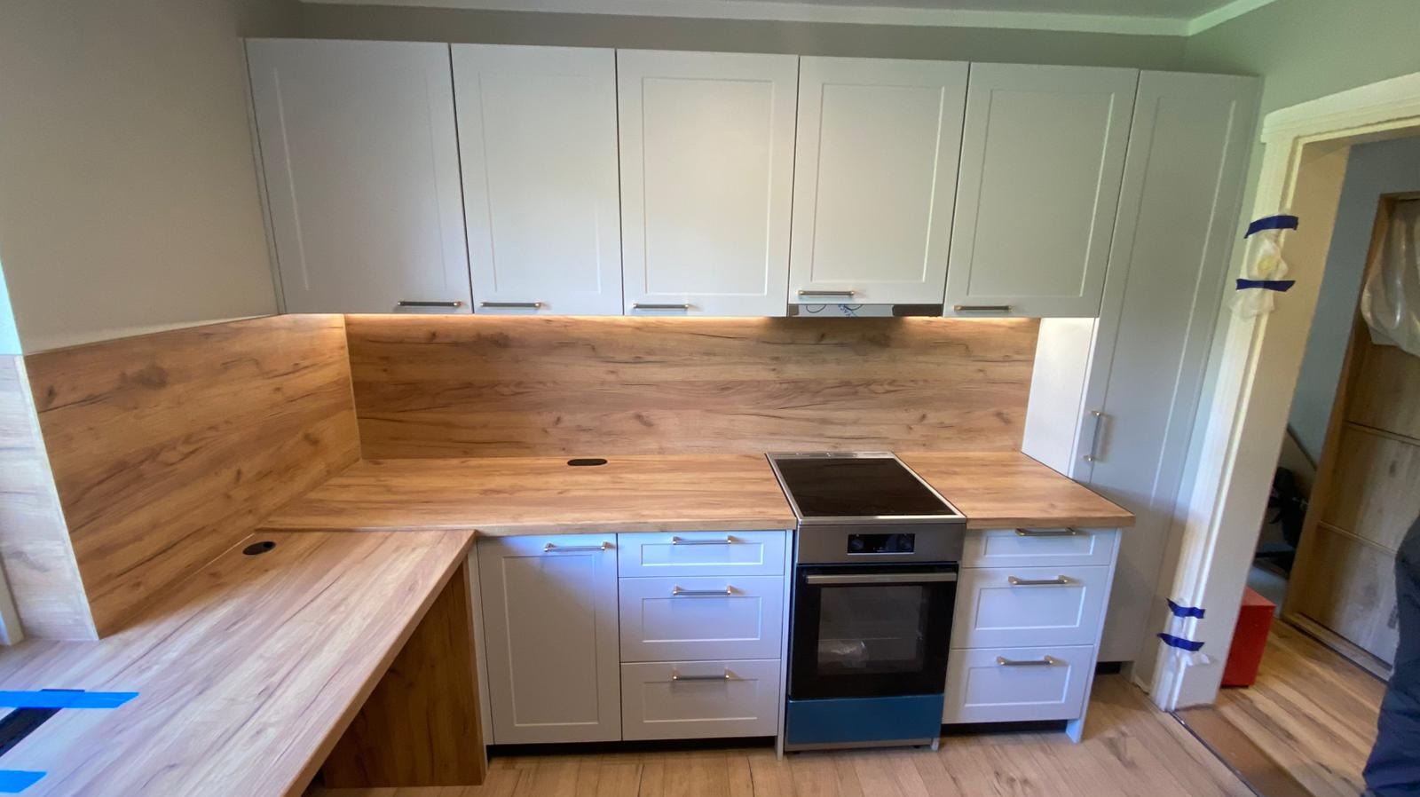 Jasna kuchnia z drewnianym blatem i oświetleniem podszafkowym. Widoczny piekarnik i płyta indukcyjna. Elementy wykończeniowe zabezpieczone folią.