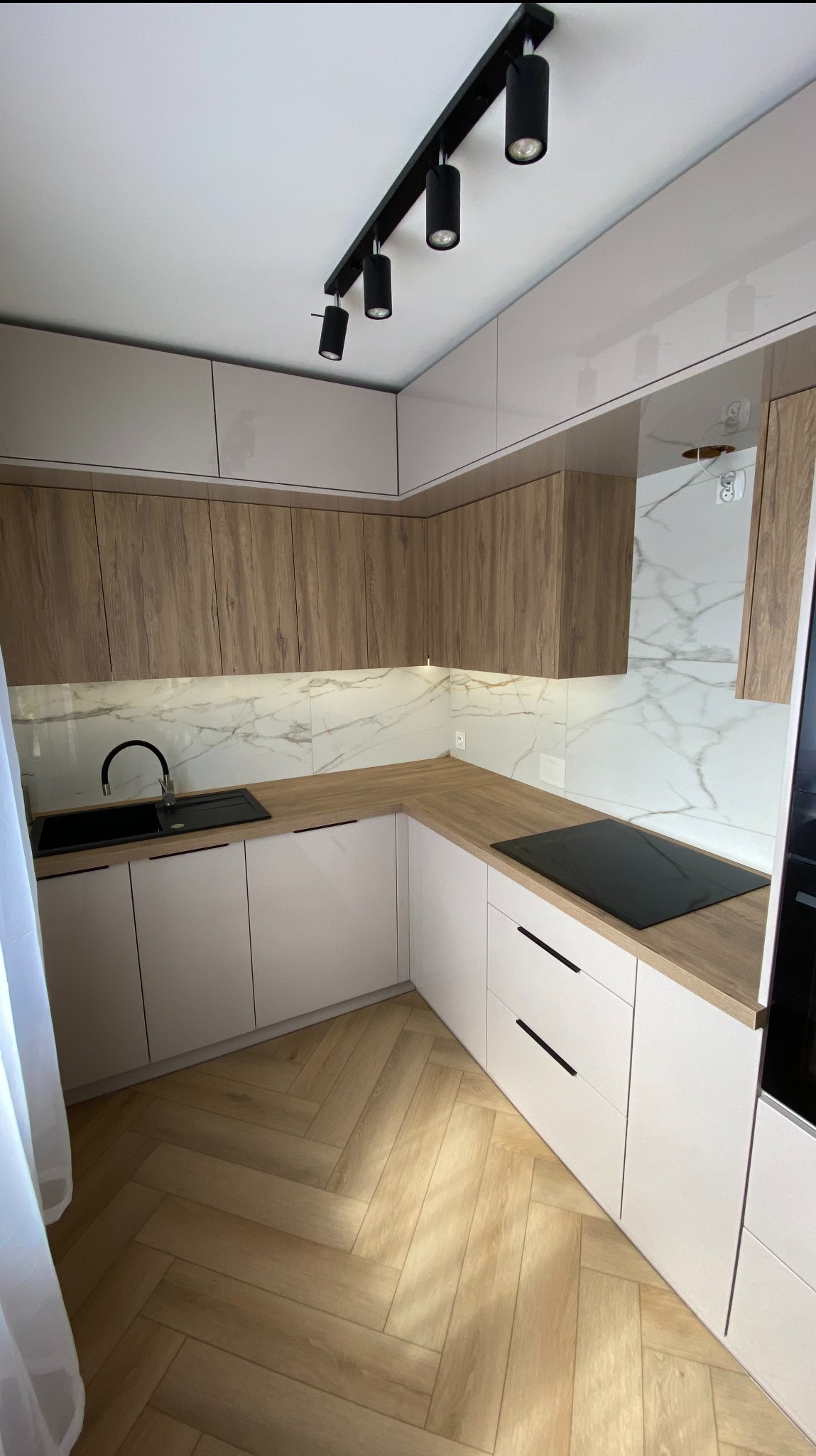 Jasna kuchnia w stylu skandynawskim: białe fronty dolne, drewnopodobne górne szafki, marmurowy backsplash, czarna armatura i oświetlenie punktowe.