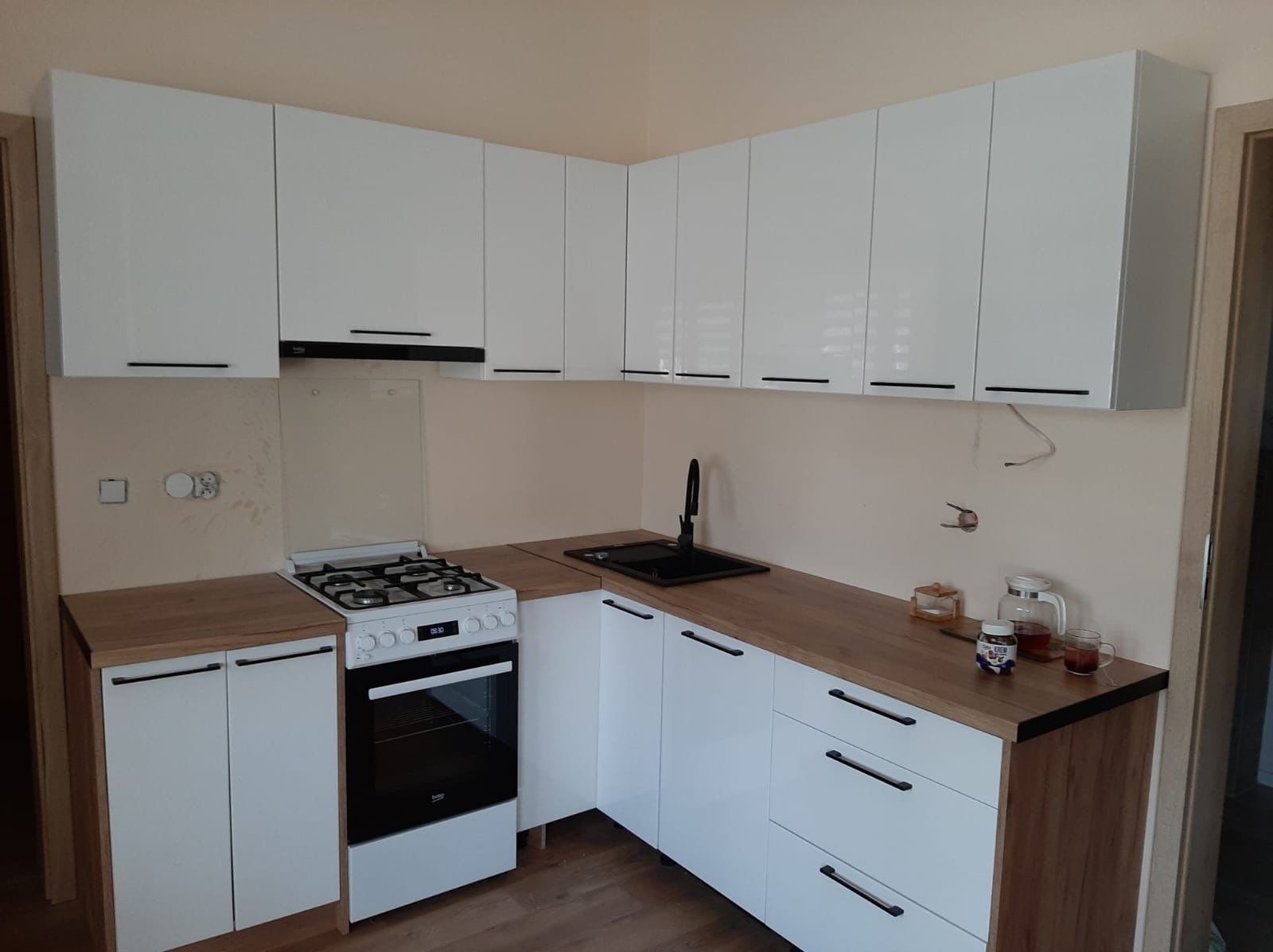 Nowoczesna kuchnia z białymi, lakierowanymi frontami i drewnianym blatem. Widoczny zlew, kuchenka i wiszące szafki z czarnymi, podłużnymi uchwytami. Minimalistyczny design.