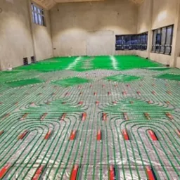 Rozłożona instalacja ogrzewania podłogowego z zielonych rurek na folii izolacyjnej w przestronnym, surowym pomieszczeniu z oknami.