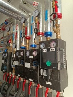 Złożony system rur i manometrów w kotłowni, z czarnymi panelami sterującymi i elementami w kolorach czerwonym i niebieskim. Widoczne złącza i oznaczenia.