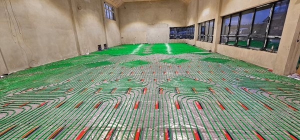 Rozłożona instalacja ogrzewania podłogowego z zielonych rurek na folii izolacyjnej w przestronnym, surowym pomieszczeniu z oknami.