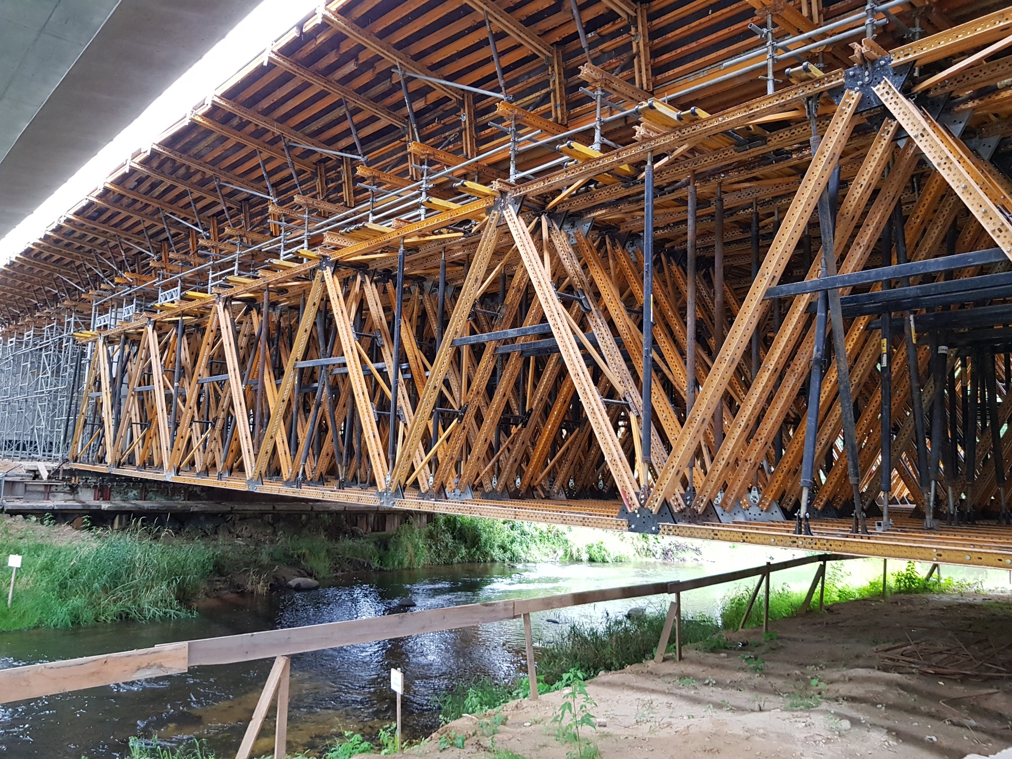 Konstrukcja wsporcza mostu w trakcie budowy nad rzeką, widok z dołu. Drewniane elementy tworzą geometryczny wzór, w tle zieleń i woda.
