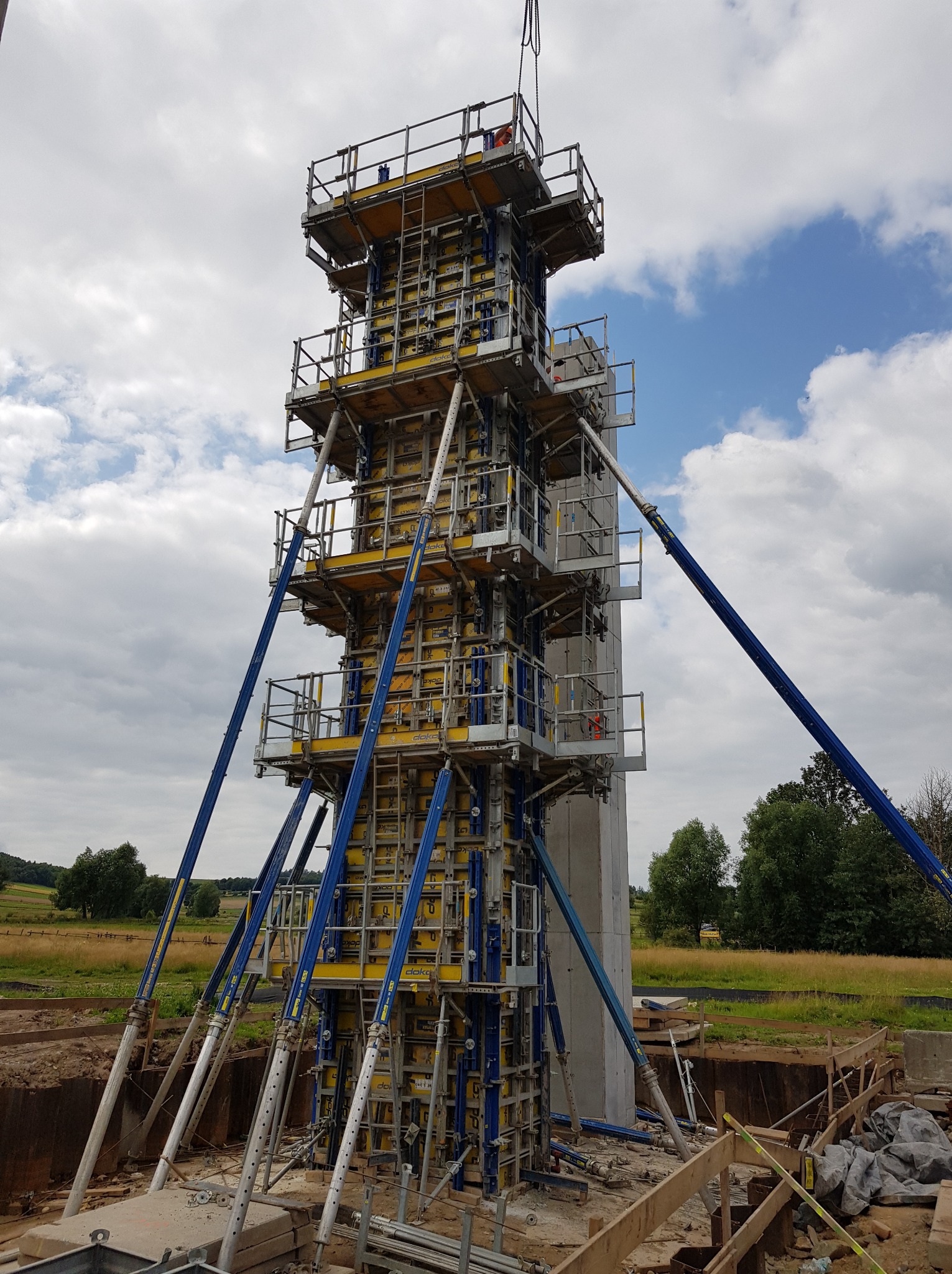 Żelbetowy słup w szalunku systemowym Doka, wsparty niebieskimi podporami, na tle krajobrazu z polami i błękitnym niebem z chmurami.