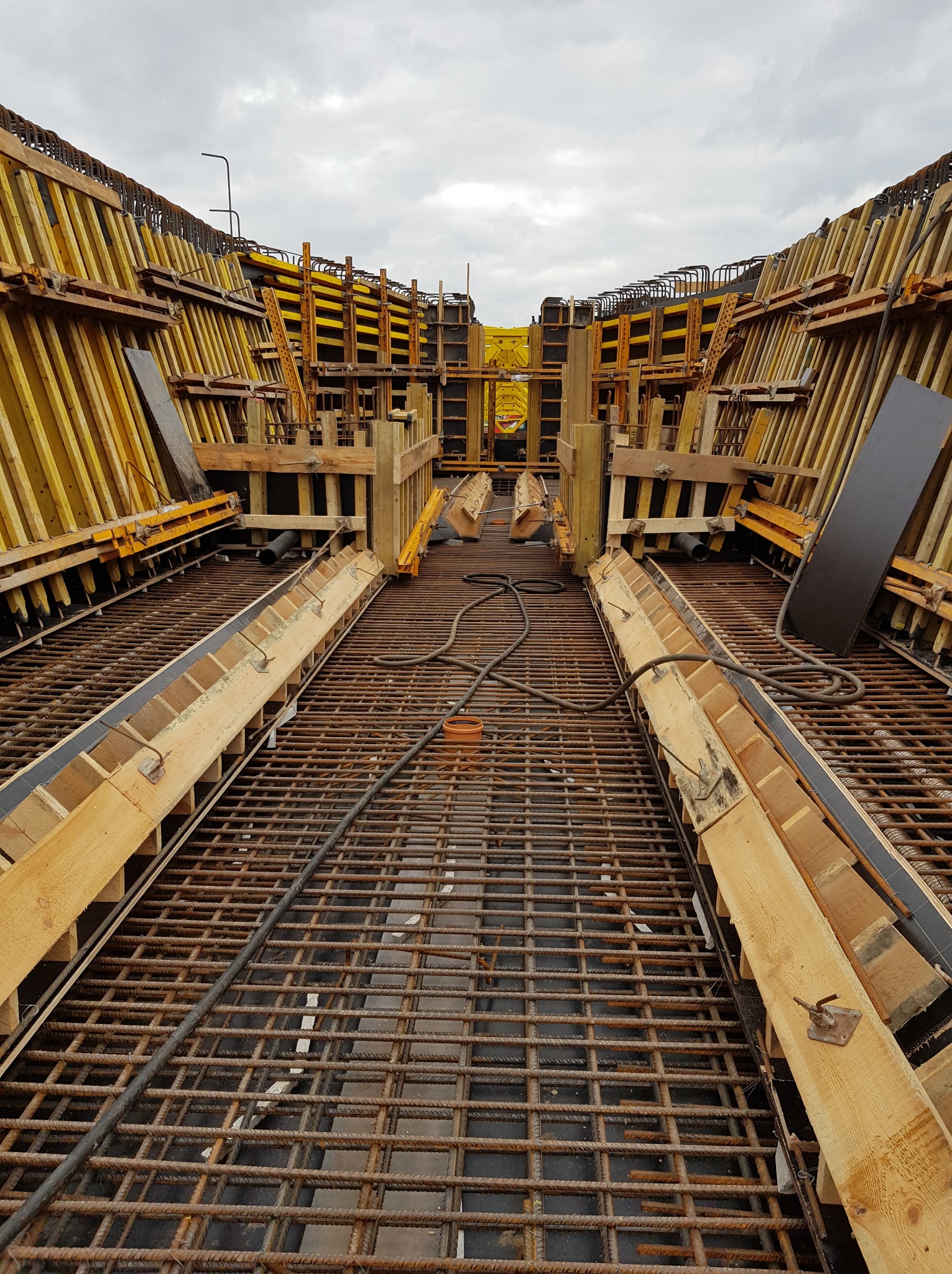Skomplikowane zbrojenie fundamentów z drewnianym szalunkiem i rurami kanalizacyjnymi, widok z góry, pochmurne niebo w tle. Precyzyjne ułożenie prętów.