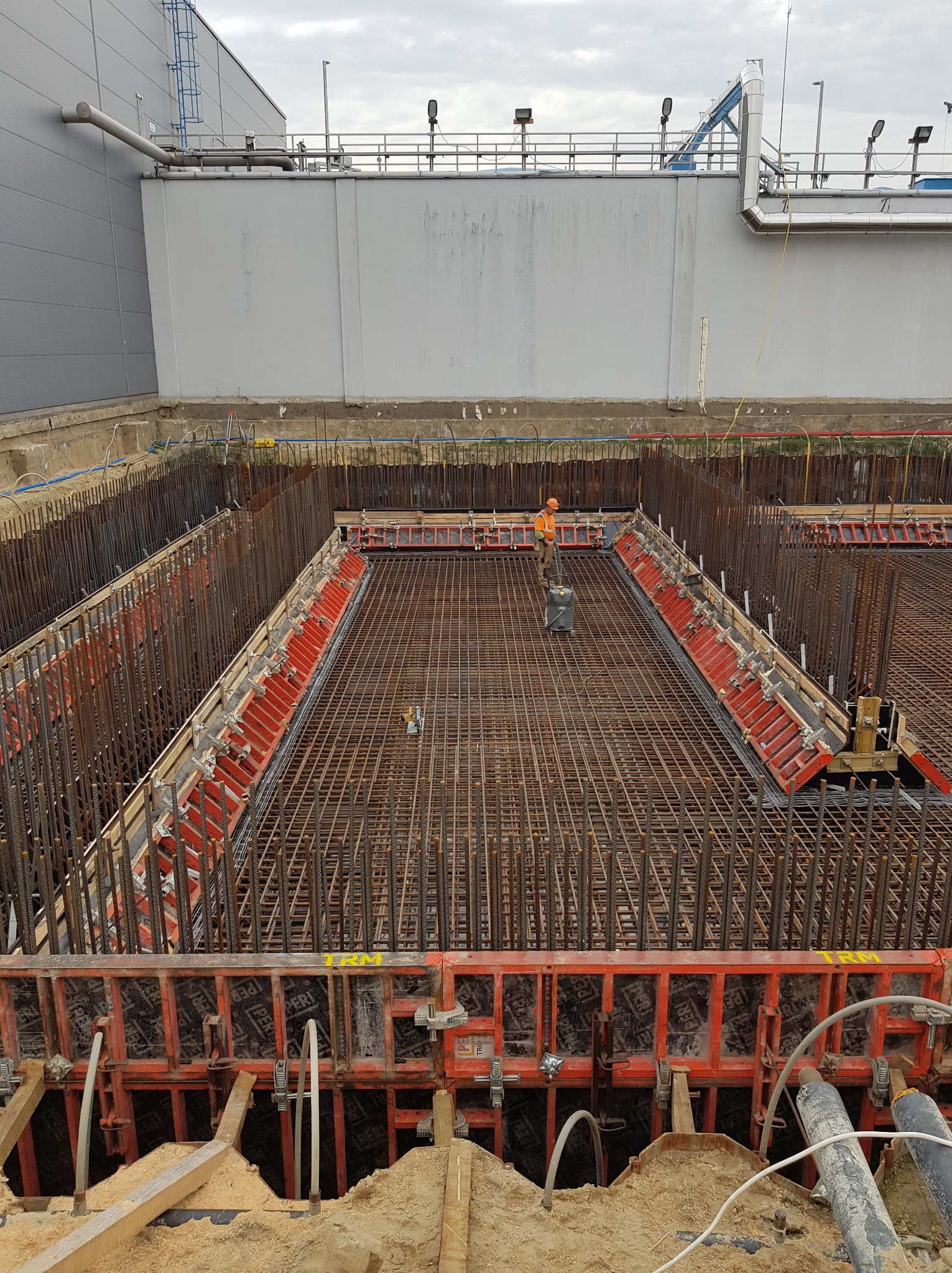 Zbrojenie fundamentów z użyciem szalunków systemowych, widok z góry na plac budowy. Pracownik w pomarańczowym ubraniu kontroluje ułożenie prętów.