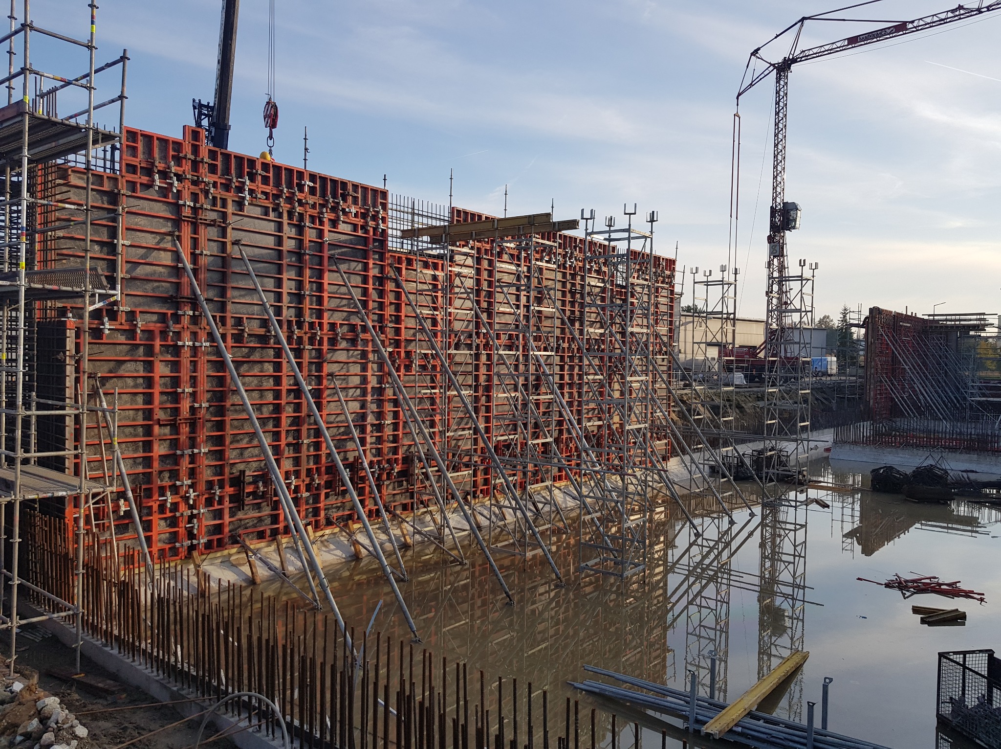 Szczegółowe ujęcie szalunków fundamentowych z widocznym zbrojeniem i stojącą wodą, wspartych rusztowaniami, z żurawiem w tle, w pochmurny dzień.