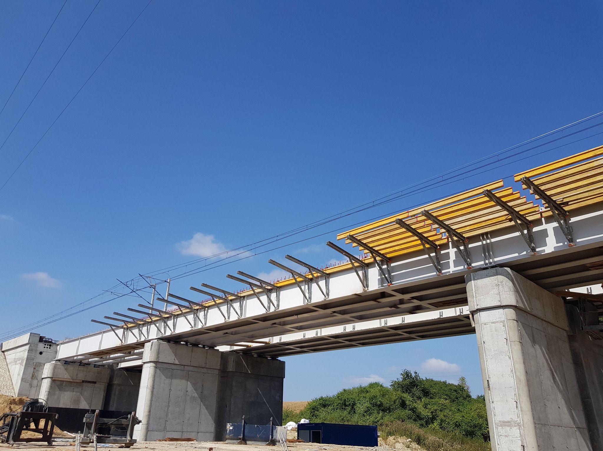 Żelbetowa konstrukcja mostu w trakcie budowy, widoczne szalunki i zbrojenie, błękitne niebo w tle. Ujęcie z dołu, podkreślające masywność konstrukcji.