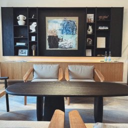 TOKON sp. z o.o. - Eleganckie wnętrze: drewniany stół, krzesła z poduszkami, szafka z dekoracjami i obrazem. Nowoczesny design z elementami artystycznymi i subtelnym oświetleniem.