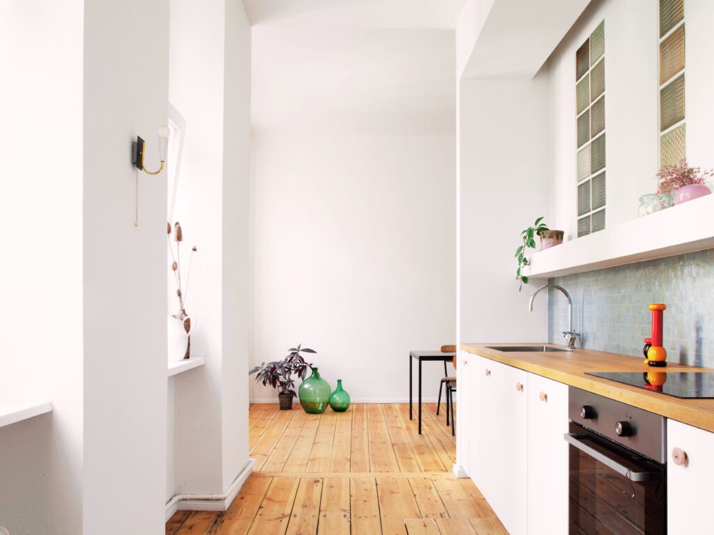 Jasna kuchnia z drewnianym blatem i podłogą, akcenty roślinne i dekoracyjne butelki. Minimalistyczny design z elementami vintage.