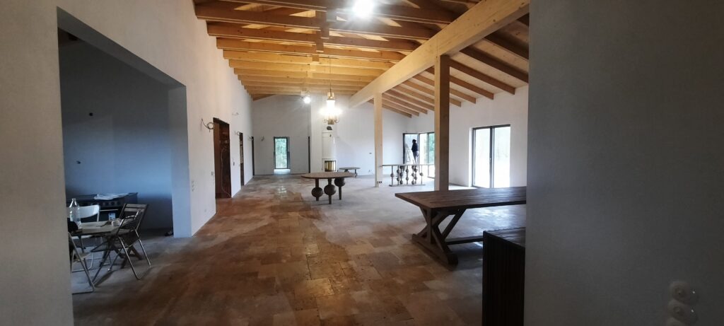 Przestronne wnętrze po remoncie z drewnianą konstrukcją dachu i jasnymi ścianami. Widoczne drewniane stoły i fragment wykończonej podłogi z płytek.