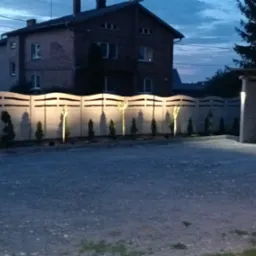 Oświetlony dekoracyjny betonowy płot z podświetlonymi drzewkami iglastymi, widok nocny. Eleganckie oświetlenie posesji wzdłuż ogrodzenia.