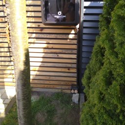 ELEKTRO-RAV RAFAŁ STASIAK - Zewnętrzna stacja ładowania pojazdów elektrycznych AmperePoint zamontowana na drewnianej elewacji budynku, obok ciemnej ściany i zielonego krzewu.