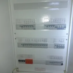 Rozdzielnia elektryczna w białej obudowie, z bezpiecznikami oznaczonymi etykietami. Widoczne przewody u dołu. Tablica z opisem obwodów.