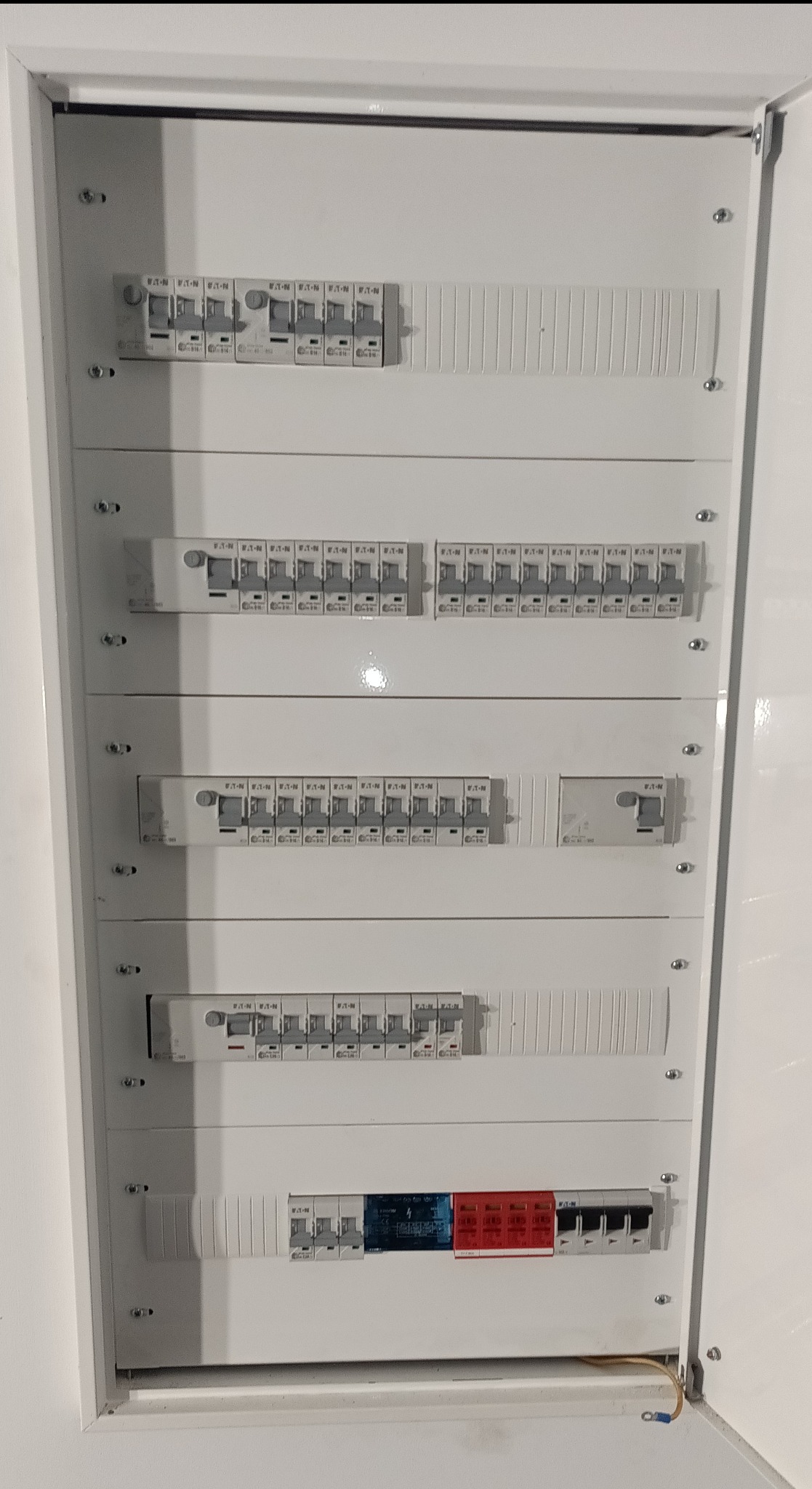 Rozdzielnica elektryczna w białej obudowie z widocznymi bezpiecznikami i wyłącznikami różnicowoprądowymi, zamontowana wewnątrz budynku.