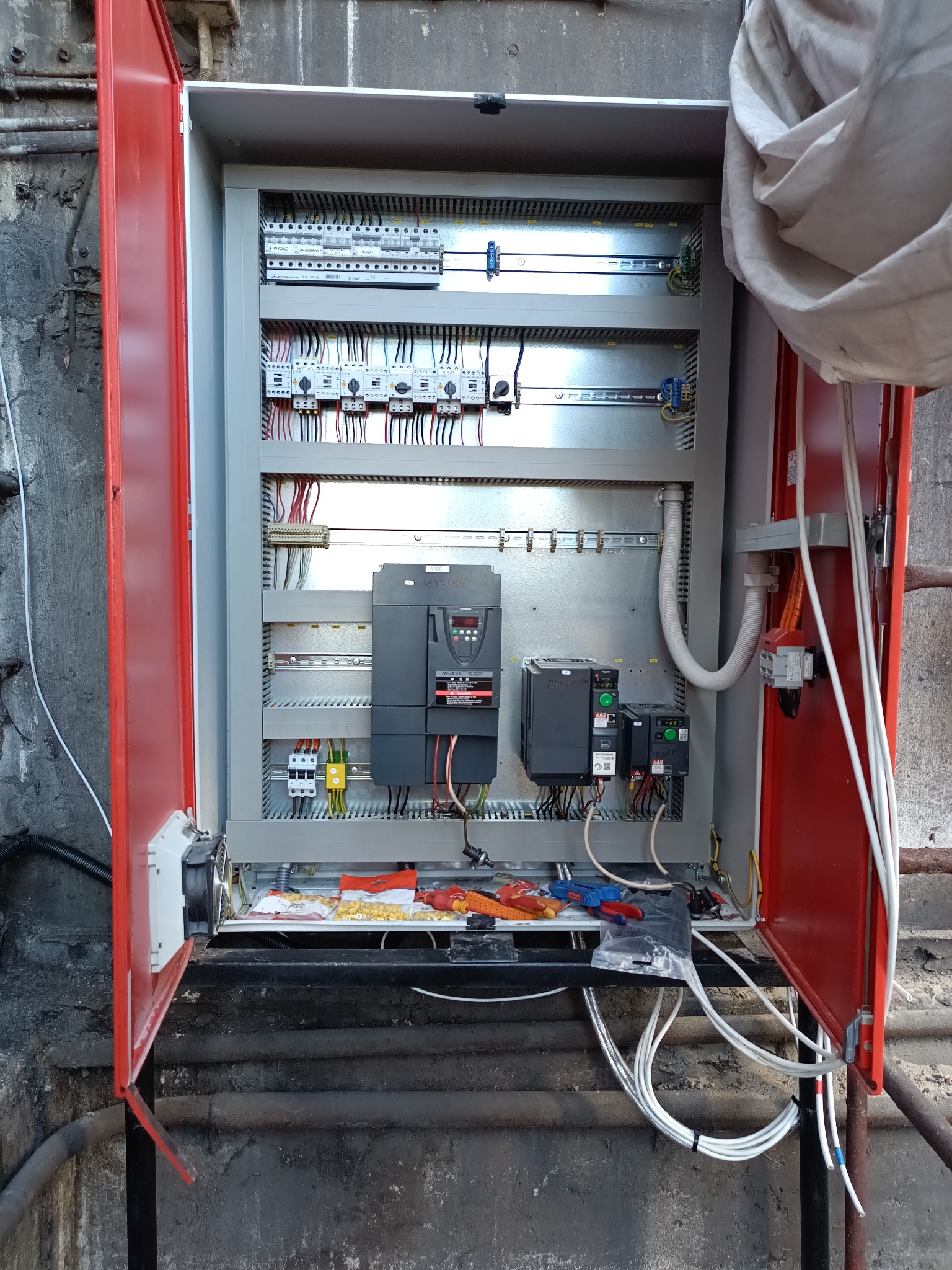 Otwarta szafa sterownicza w kolorze czerwonym, z widocznymi elementami elektrycznymi wewnątrz, takimi jak przekaźniki i okablowanie. Widoczne narzędzia w dolnej części szafy.