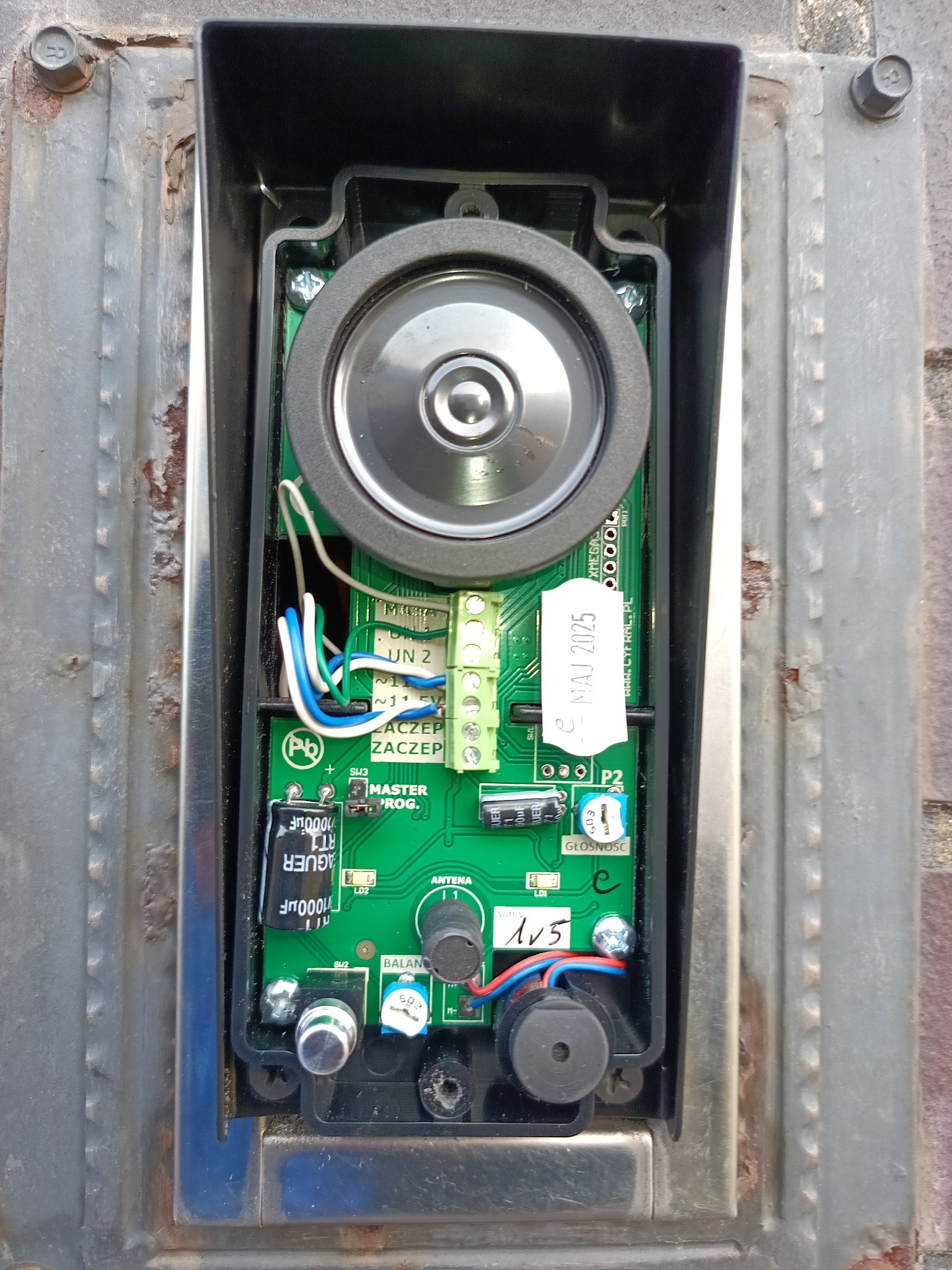 Otwarta obudowa domofonu/wideofonu z widoczną płytką drukowaną, głośnikiem i okablowaniem. Widoczne elementy elektroniczne i naklejka z datą ważności. Metalowa obudowa.