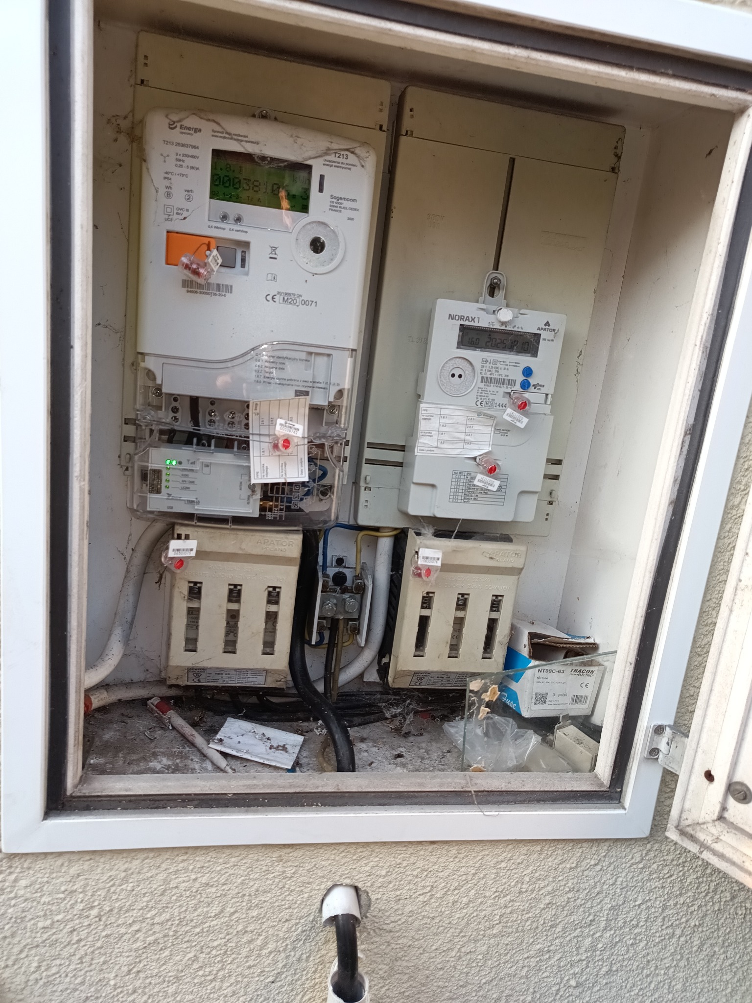 Otwarta skrzynka z dwoma licznikami energii elektrycznej i bezpiecznikami. Widoczne kable, elementy instalacji i obudowa skrzynki z zewnątrz.