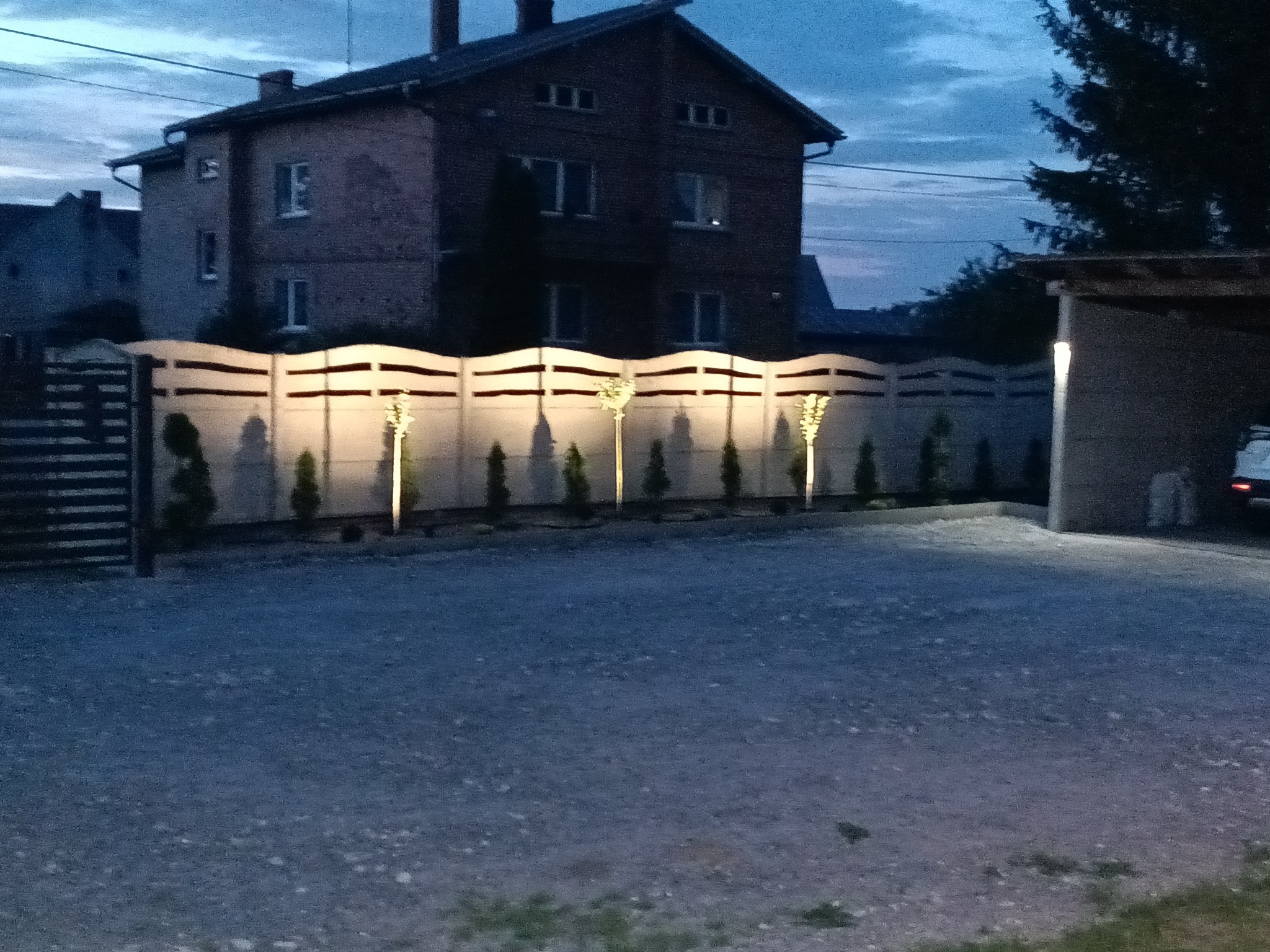 Oświetlony dekoracyjny betonowy płot z podświetlonymi drzewkami iglastymi, widok nocny. Eleganckie oświetlenie posesji wzdłuż ogrodzenia.