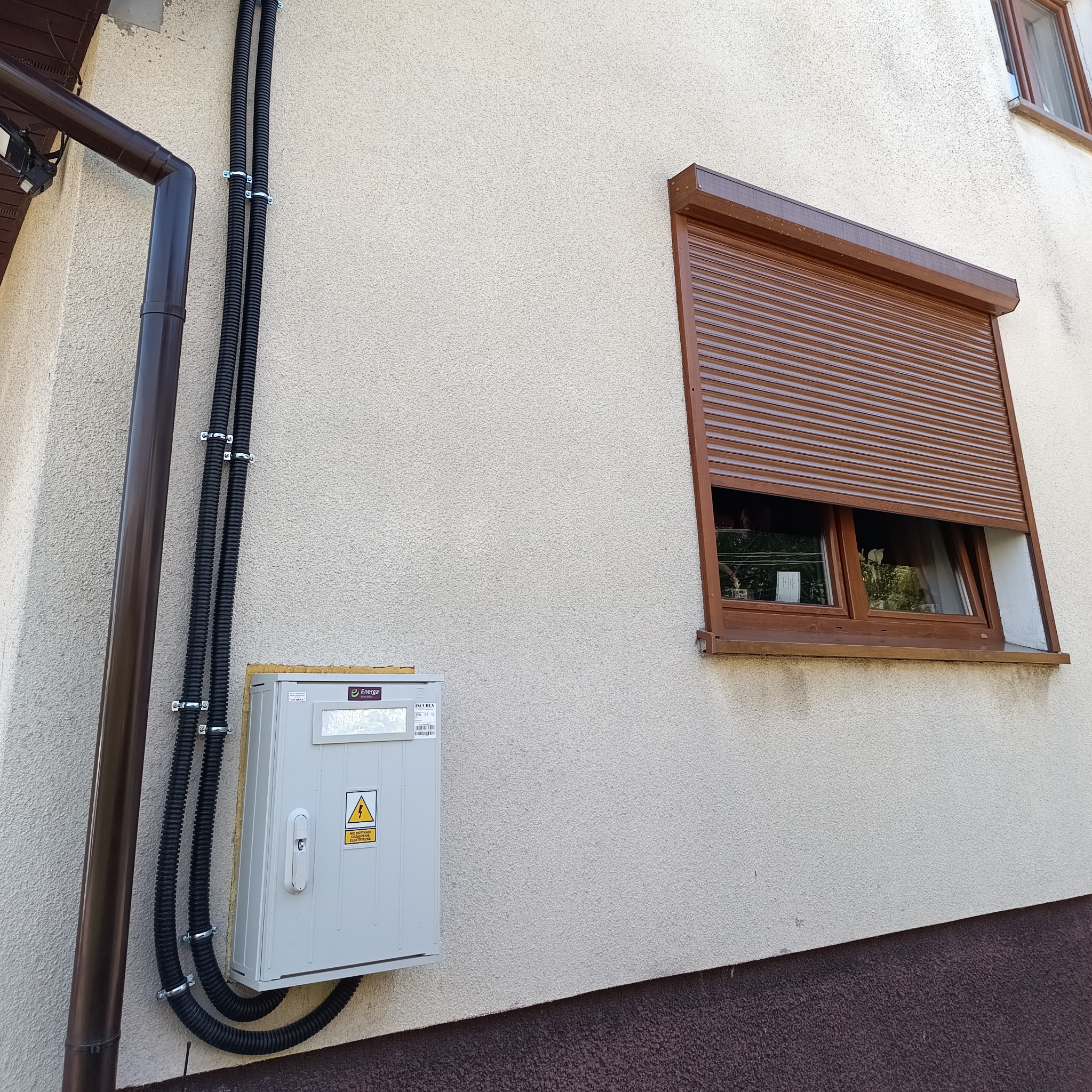 Zewnętrzna skrzynka elektryczna Energa z żółtym znakiem ostrzegawczym. Dwie czarne rury ochronne prowadzą wzdłuż ściany. Obok brązowa rynna i okno z brązową roletą.