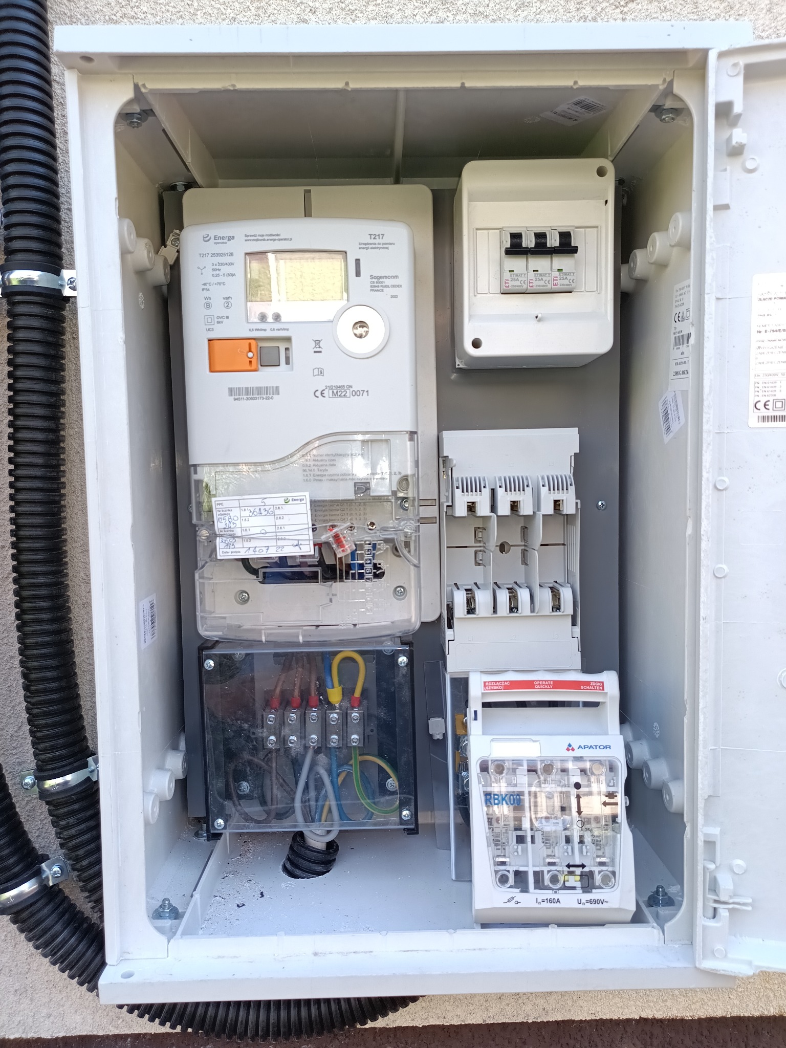 Biała skrzynka elektryczna z licznikiem, bezpiecznikami i przewodami. Widoczne czarne rury karbowane doprowadzające kable. Instalacja na zewnątrz budynku.