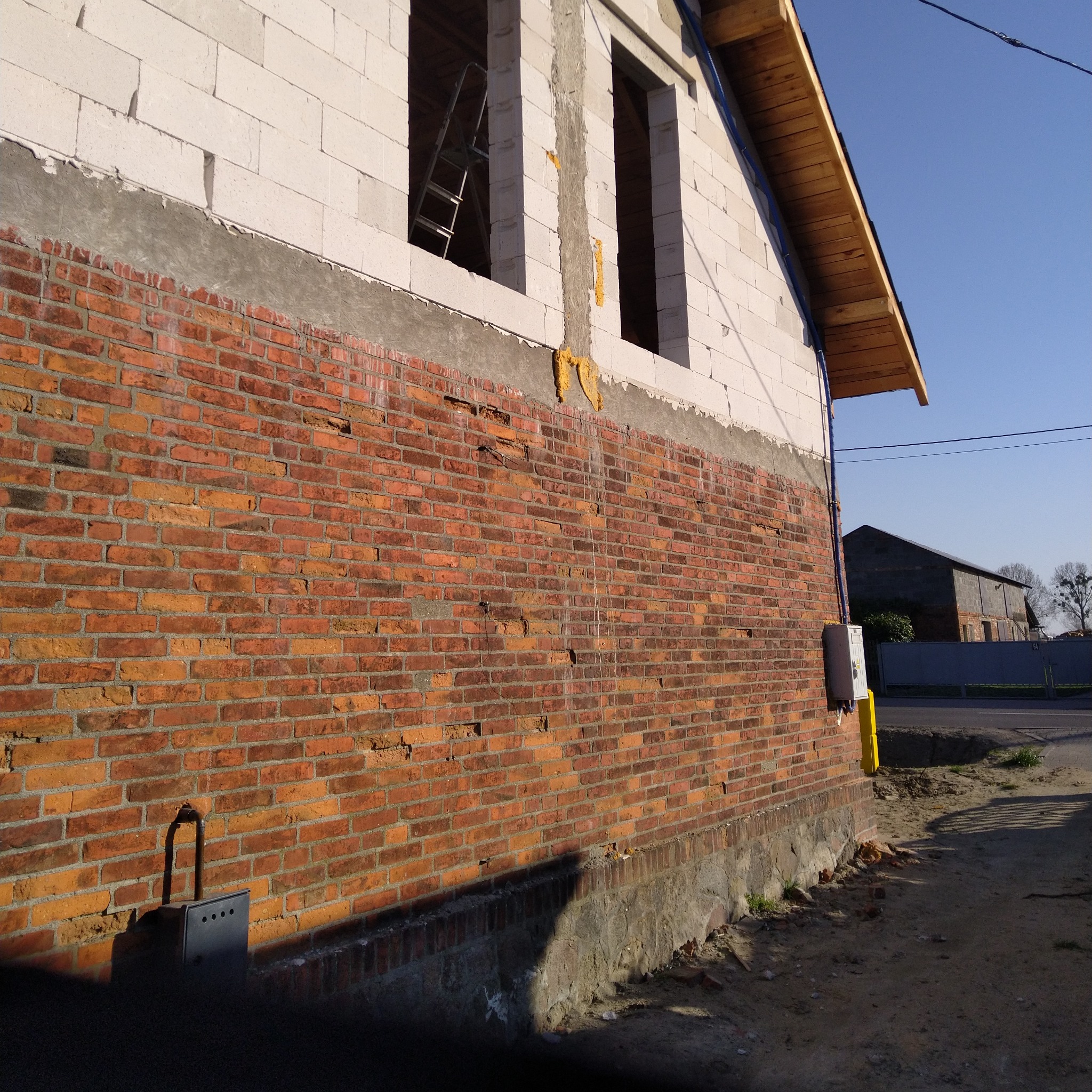 Ceglany mur budynku w trakcie budowy, z widoczną instalacją elektryczną i pustakami na górze. W tle błękitne niebo i budynek gospodarczy.