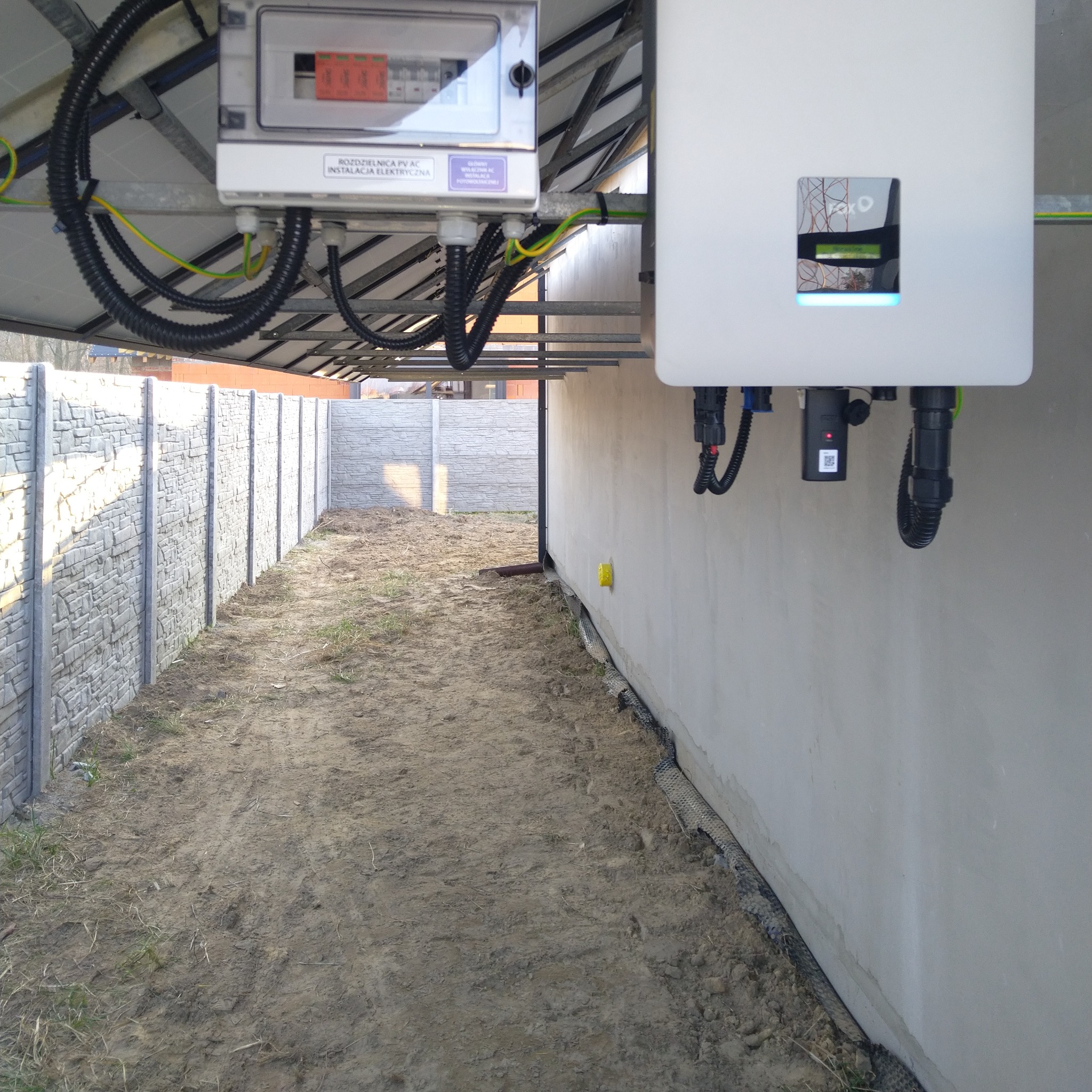 Rozdzielnia PV AC i falownik Fronius na zewnątrz budynku, widoczne kable w peszlach. Szara, betonowa ściana i ogrodzenie panelowe w tle. Instalacja elektryczna fotowoltaiczna.