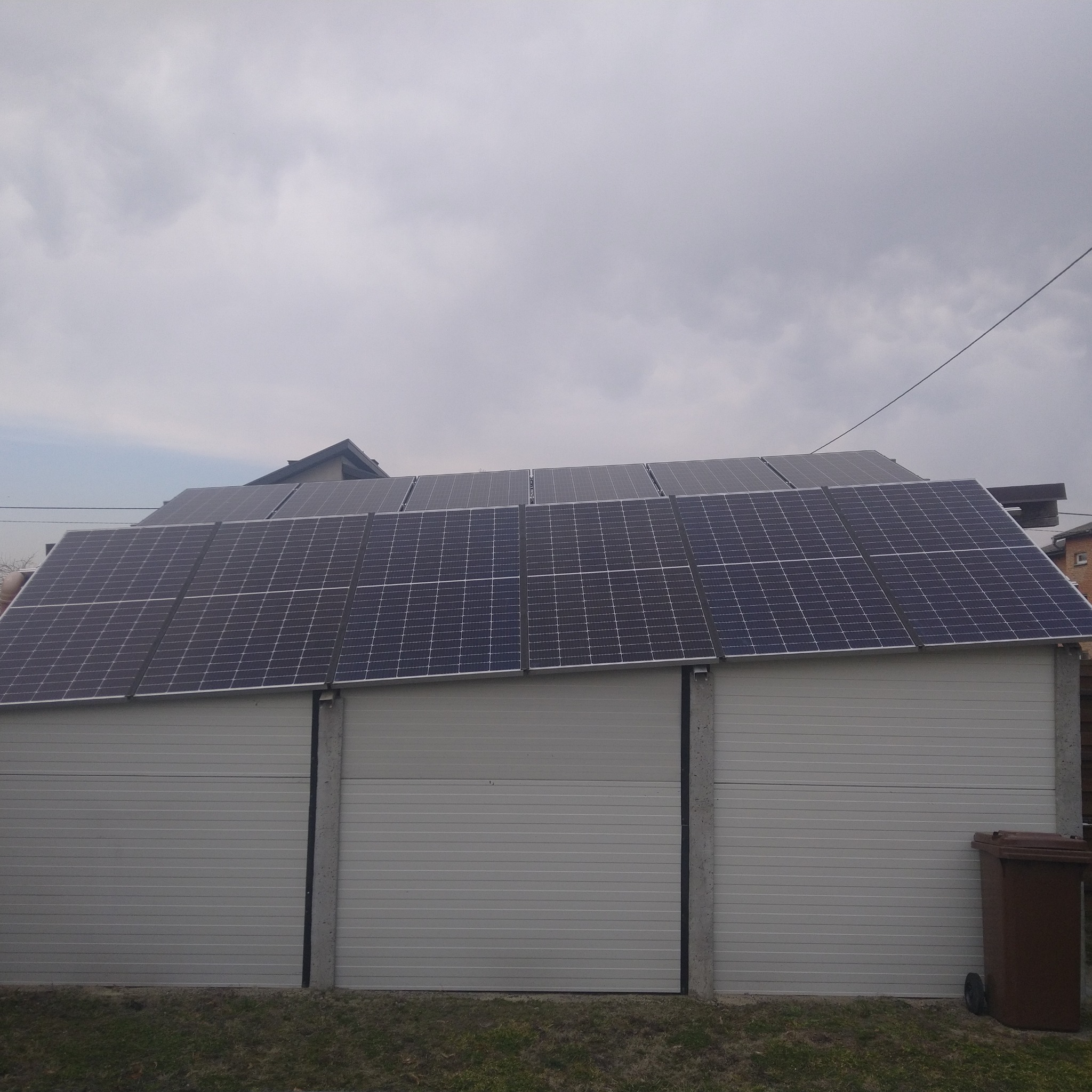 Panele słoneczne na dachu garażu z segmentową bramą w pochmurny dzień. Widoczny brązowy kosz na śmieci.