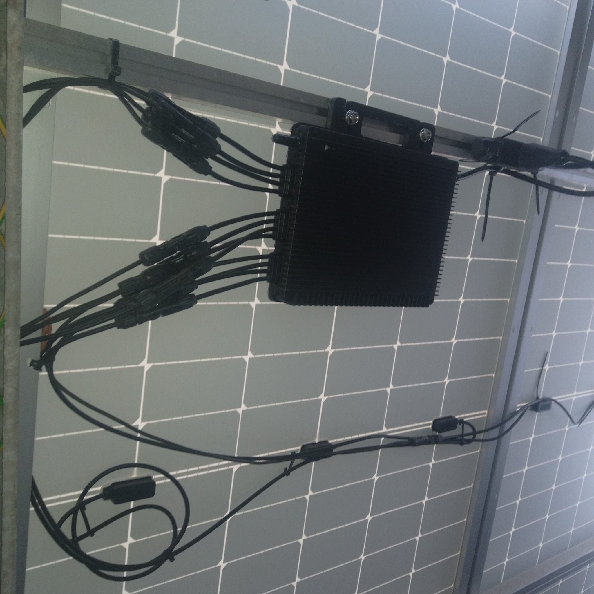 Zamontowany inwerter solarny z przewodami na panelach fotowoltaicznych. Widoczne złącza kablowe i metalowa konstrukcja montażowa. Kątowe ujęcie.