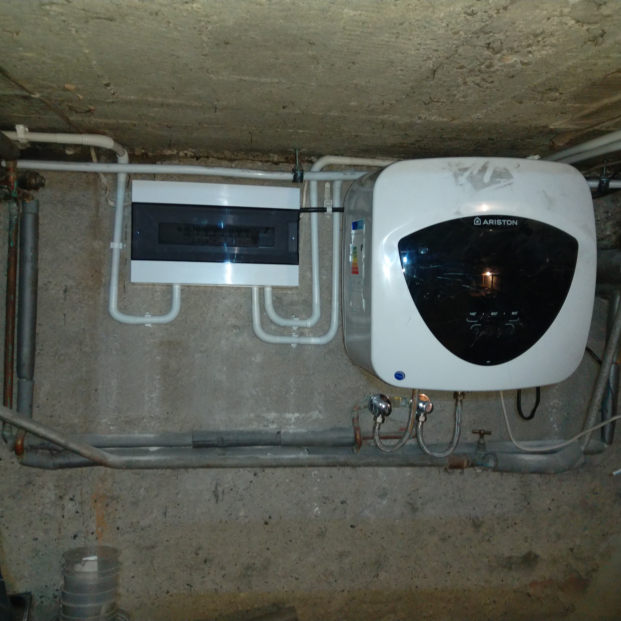 Biały podgrzewacz wody Ariston, zamontowany obok skrzynki bezpiecznikowej i rur na szarej, betonowej ścianie. Widoczne zawory i przewody zasilające.