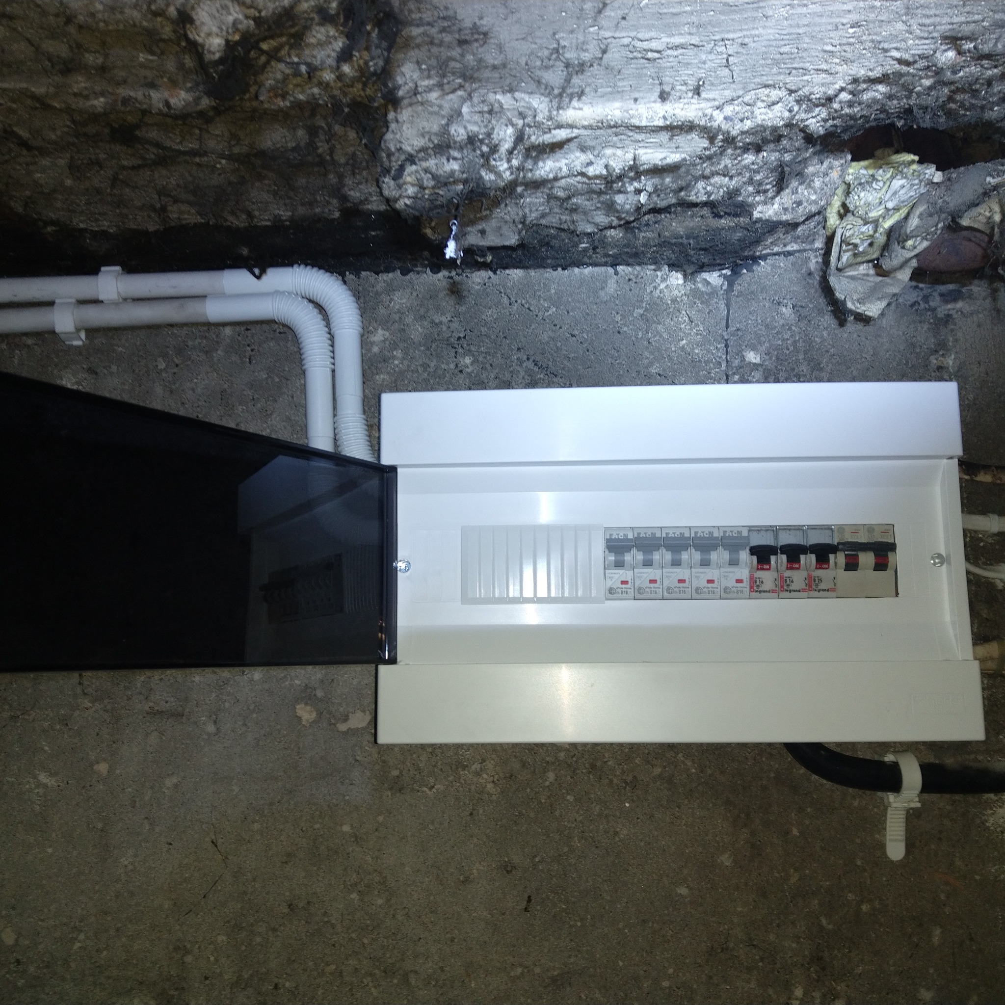 Zamontowana, biała skrzynka rozdzielcza z czarną, uchyloną osłoną, umieszczona na szarej, betonowej ścianie. Nad nią widoczne rury instalacyjne i surowy strop.