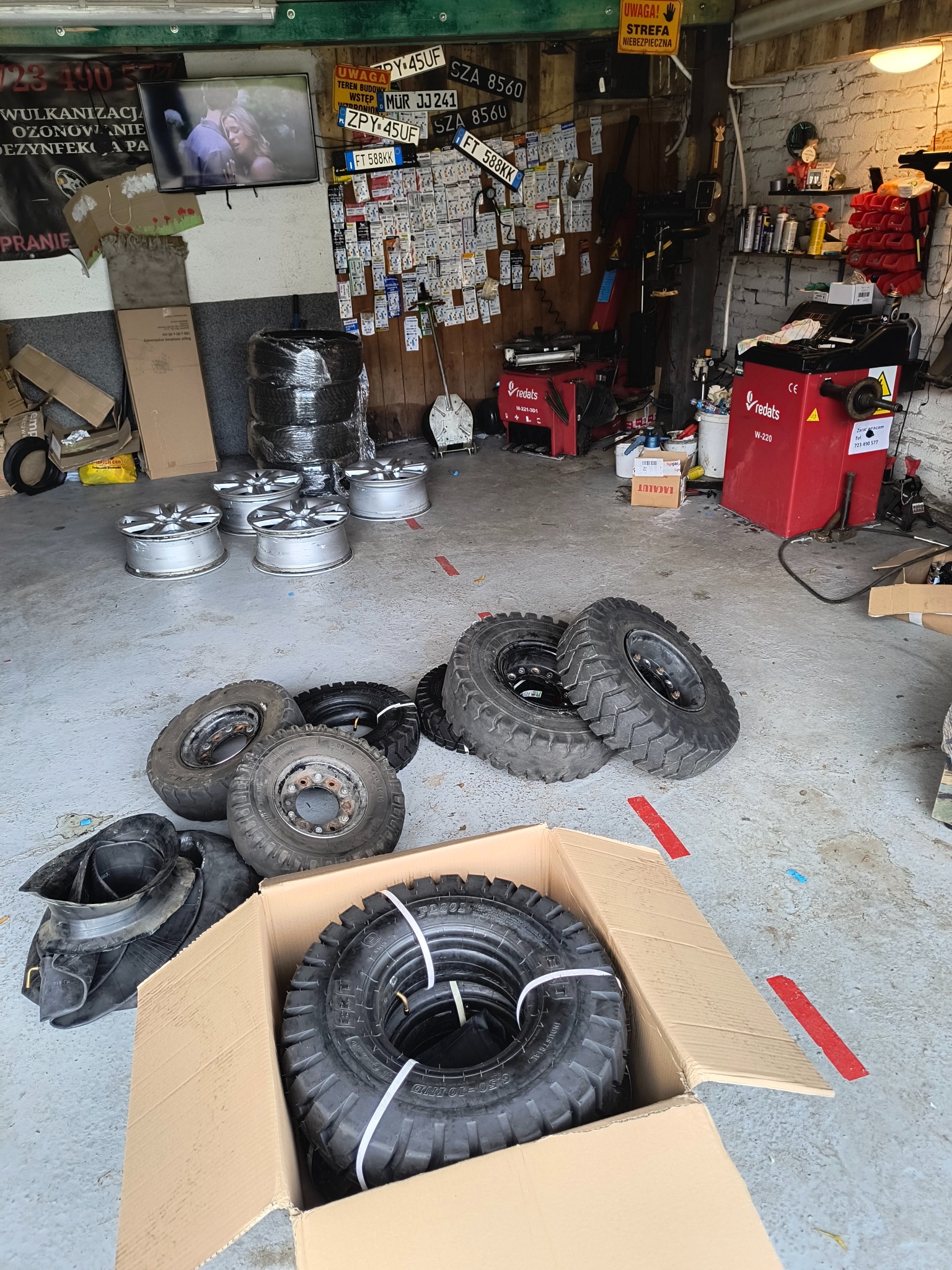 Wnętrze warsztatu z oponami w kartonie, felgami i urządzeniami do wyważania kół. Na ścianie tablice rejestracyjne i telewizor.