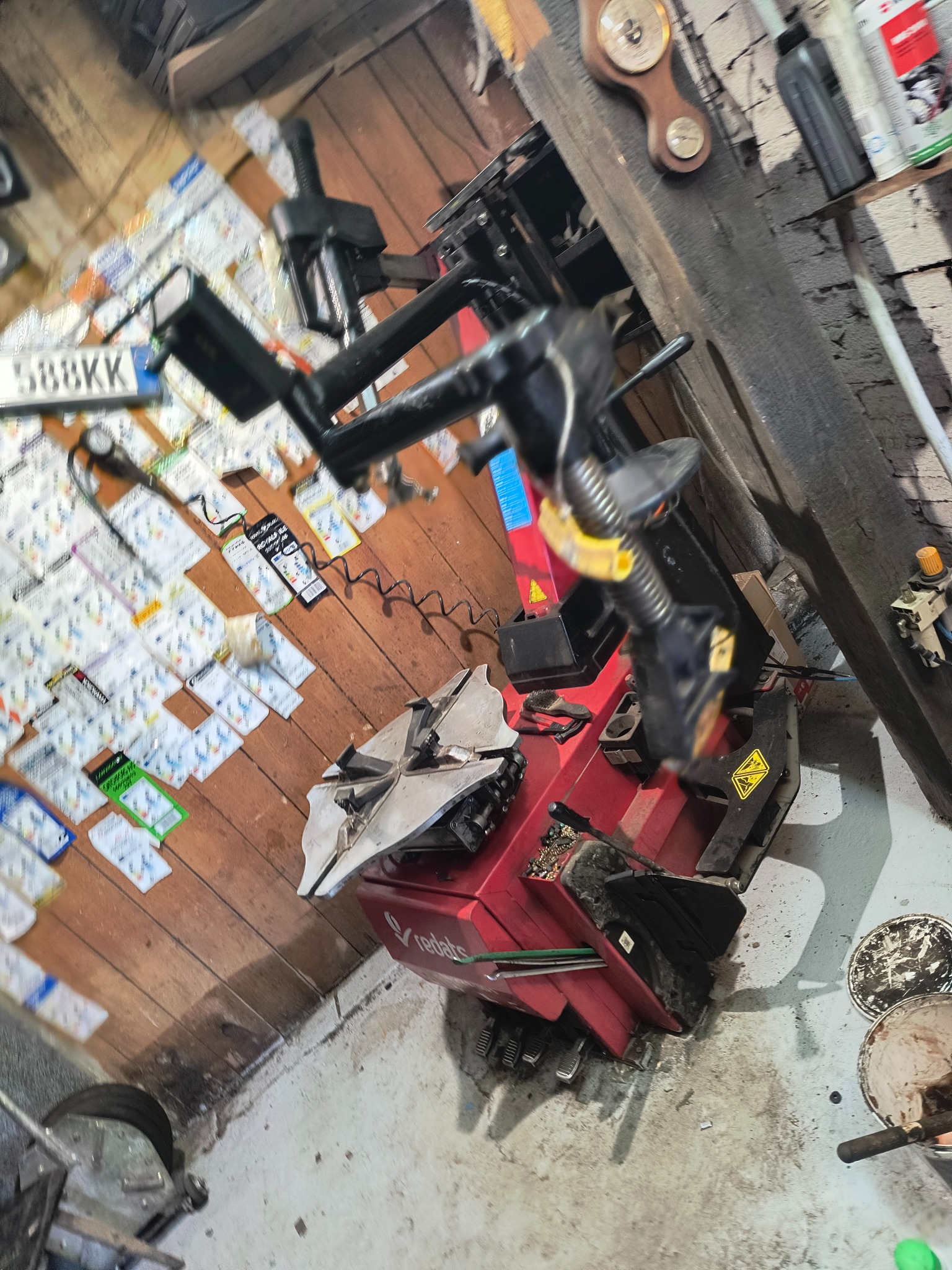 Czerwona montażownica do opon Redats w warsztacie, z widoczną ścianą z ulotkami i tablicą rejestracyjną. Sprzęt warsztatowy w nieprofesjonalnym otoczeniu.