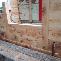 Usługi Stolarskie Rafał Gnat - Wnętrze drewnianej konstrukcji w trakcie budowy; widoczne deski, fragment starej ściany z tapetą i czerwony element ramy okiennej. Surowy stan, etap prac wykończeniowych.