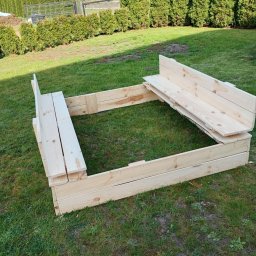 Usługi Stolarskie Rafał Gnat - Drewniana piaskownica z ławeczkami, wykonana z jasnego drewna, ustawiona na trawniku w ogrodzie. Widoczne elementy konstrukcyjne i naturalna faktura drewna.