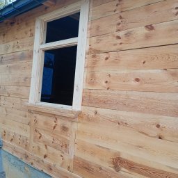 Usługi Stolarskie Rafał Gnat - Ściana budynku obita jasnym drewnem z zamontowanym oknem bez szyby. Widoczne napisy ołówkiem na desce. Surowy stan wykończenia.