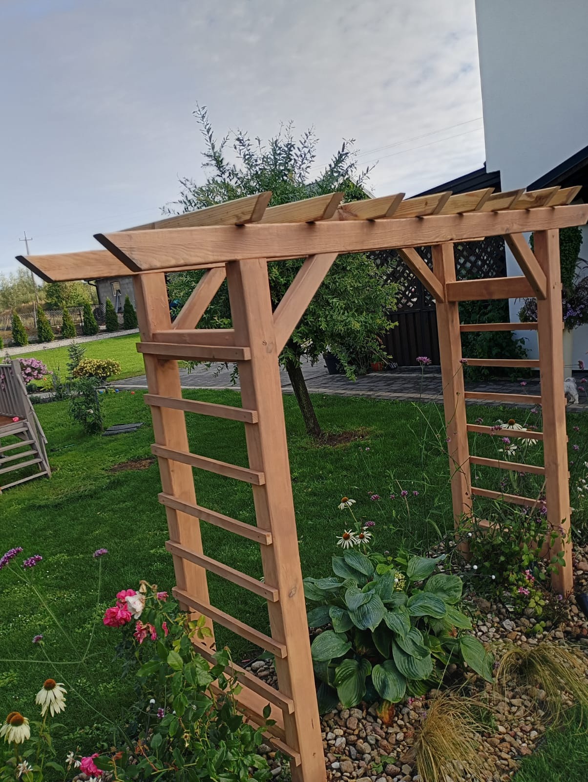 Drewniana pergola z drabinkami w ogrodzie, otoczona kwiatami i zielenią. Konstrukcja z jasnego drewna, widok na trawnik i dom w tle. Słoneczny dzień.