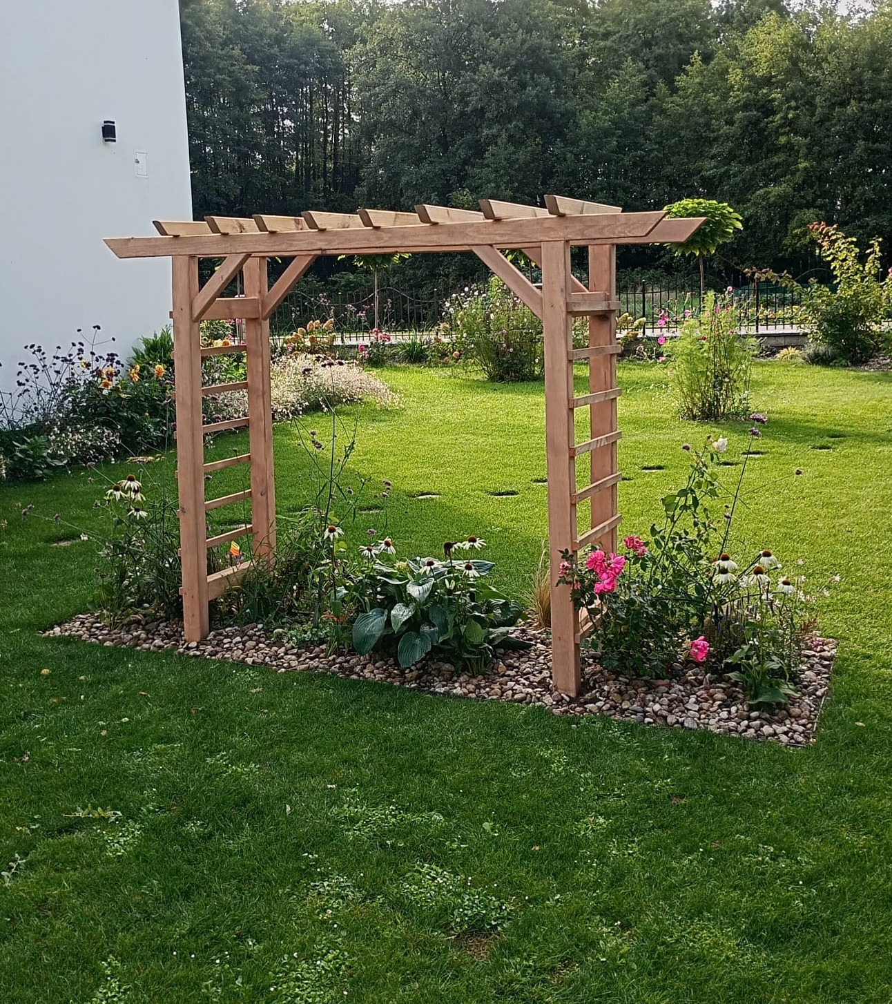 Naturalna pergola z jasnego drewna w ogrodzie, otoczona kwiatami i kamieniami, na tle zielonego trawnika i drzew. Widok z przodu, w słoneczny dzień.