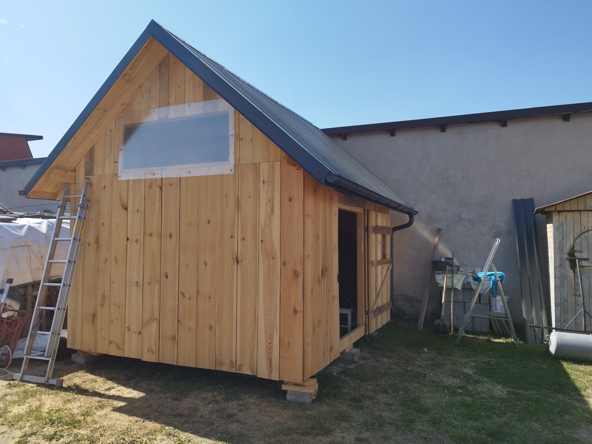 Surowa, drewniana altana z czarnym dachem, częściowo ukończona, z oknem z pleksi i drabiną opartą o ścianę. Widoczne narzędzia i materiały budowlane.