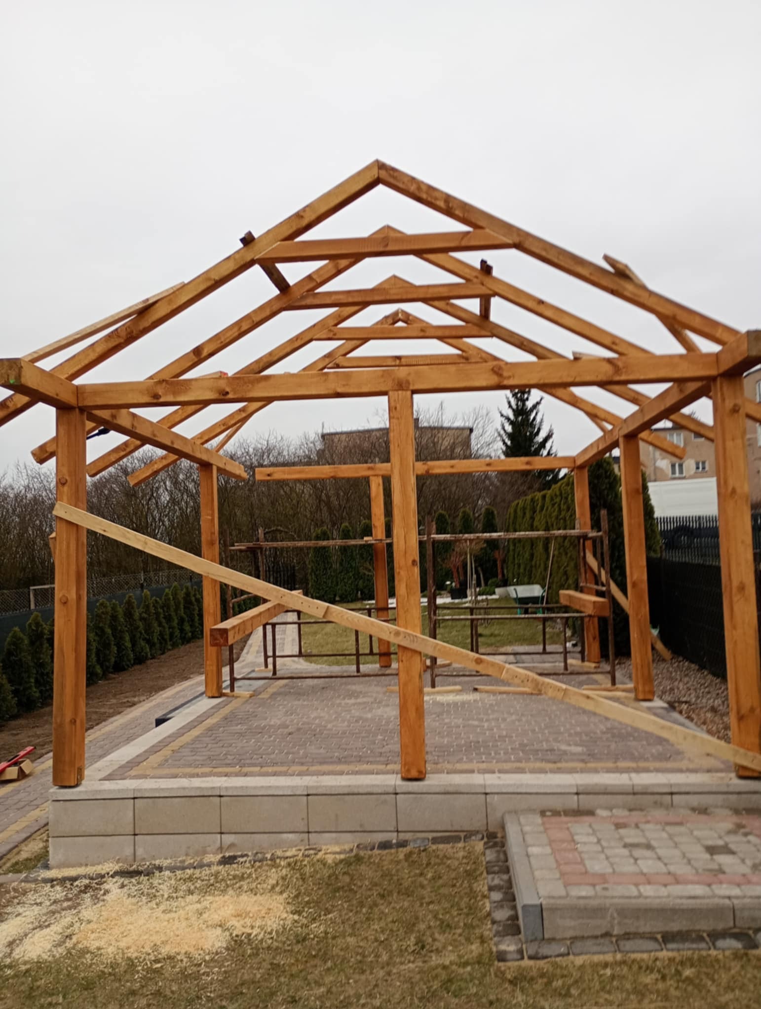 Konstrukcja drewnianej altany w trakcie budowy, widoczne elementy szkieletu, podłoże z kostki brukowej, w tle zieleń i budynki.