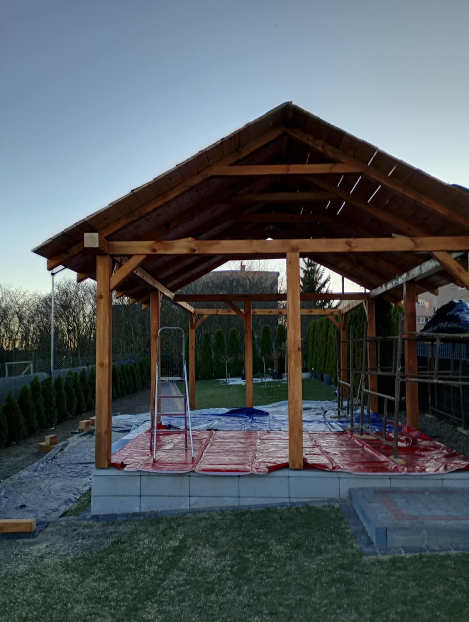 Drewniana altana w trakcie budowy, widoczne rusztowania i zabezpieczenia podłoża. Konstrukcja z belek na fundamencie, w tle drzewa i błękitne niebo o zmierzchu.