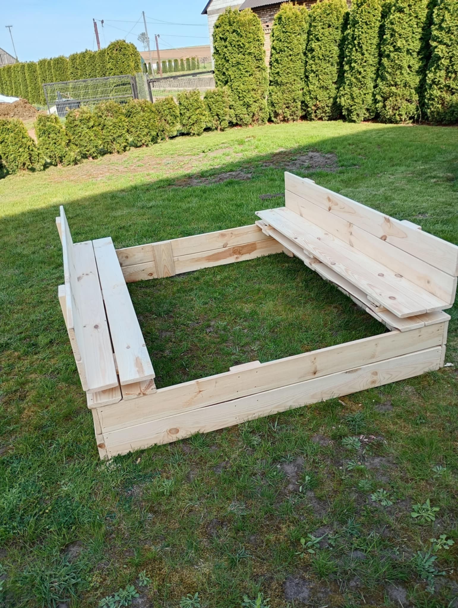 Drewniana piaskownica z ławeczkami, wykonana z jasnego drewna, ustawiona na trawniku w ogrodzie. Widoczne elementy konstrukcyjne i naturalna faktura drewna.