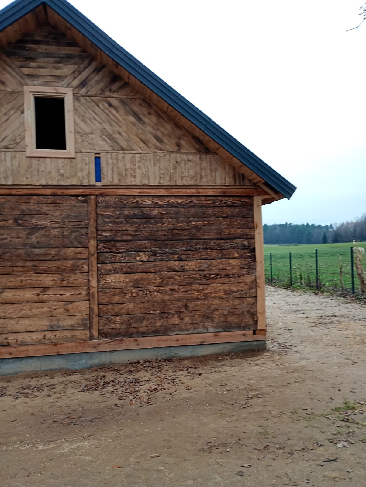 Drewniana konstrukcja budynku z ciemnym dachem i oknem bez szyby, widok z boku na tle zielonego pola i drzew. Elementy wykończeniowe w surowym drewnie.