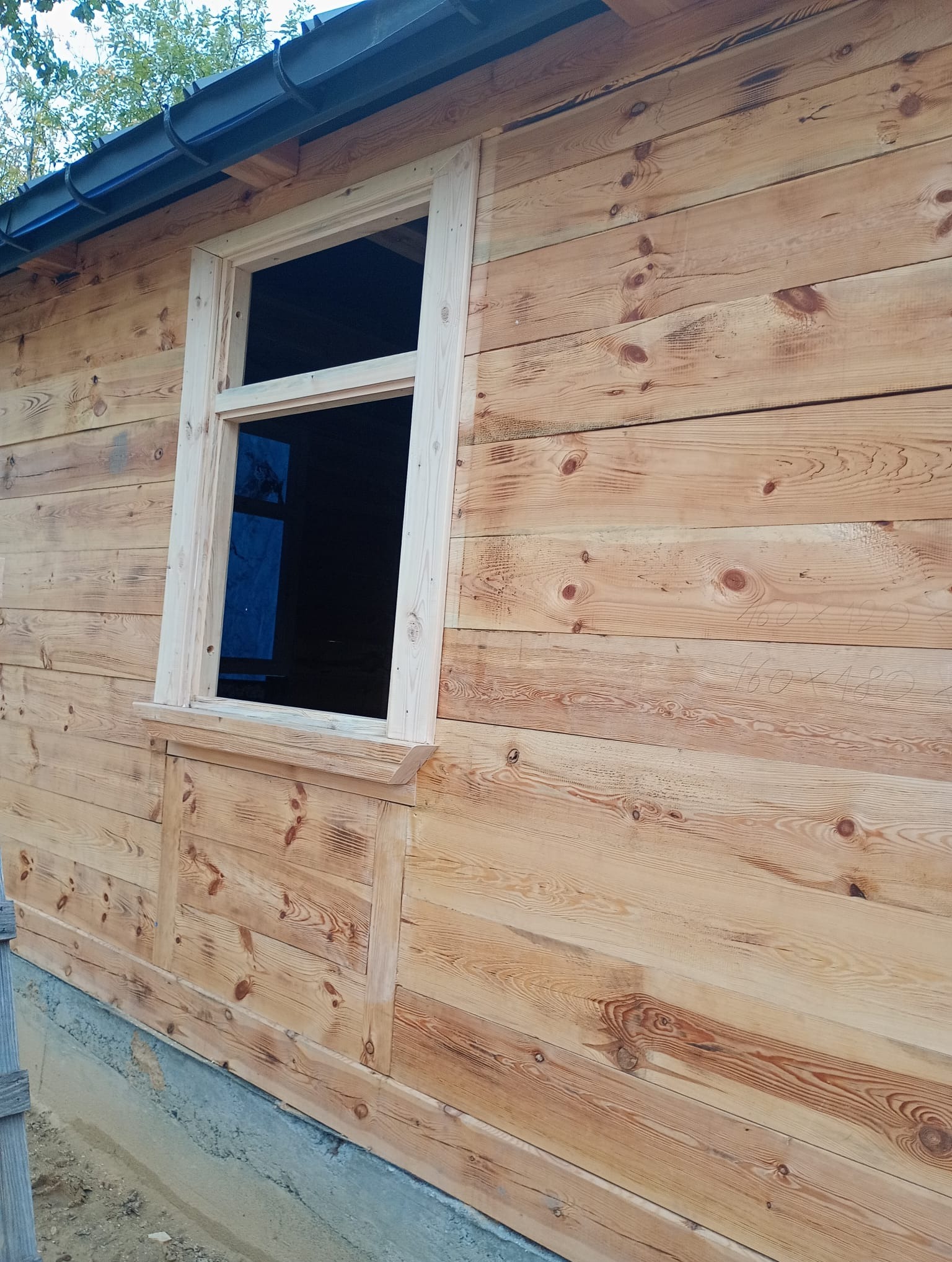 Ściana budynku obita jasnym drewnem z zamontowanym oknem bez szyby. Widoczne napisy ołówkiem na desce. Surowy stan wykończenia.