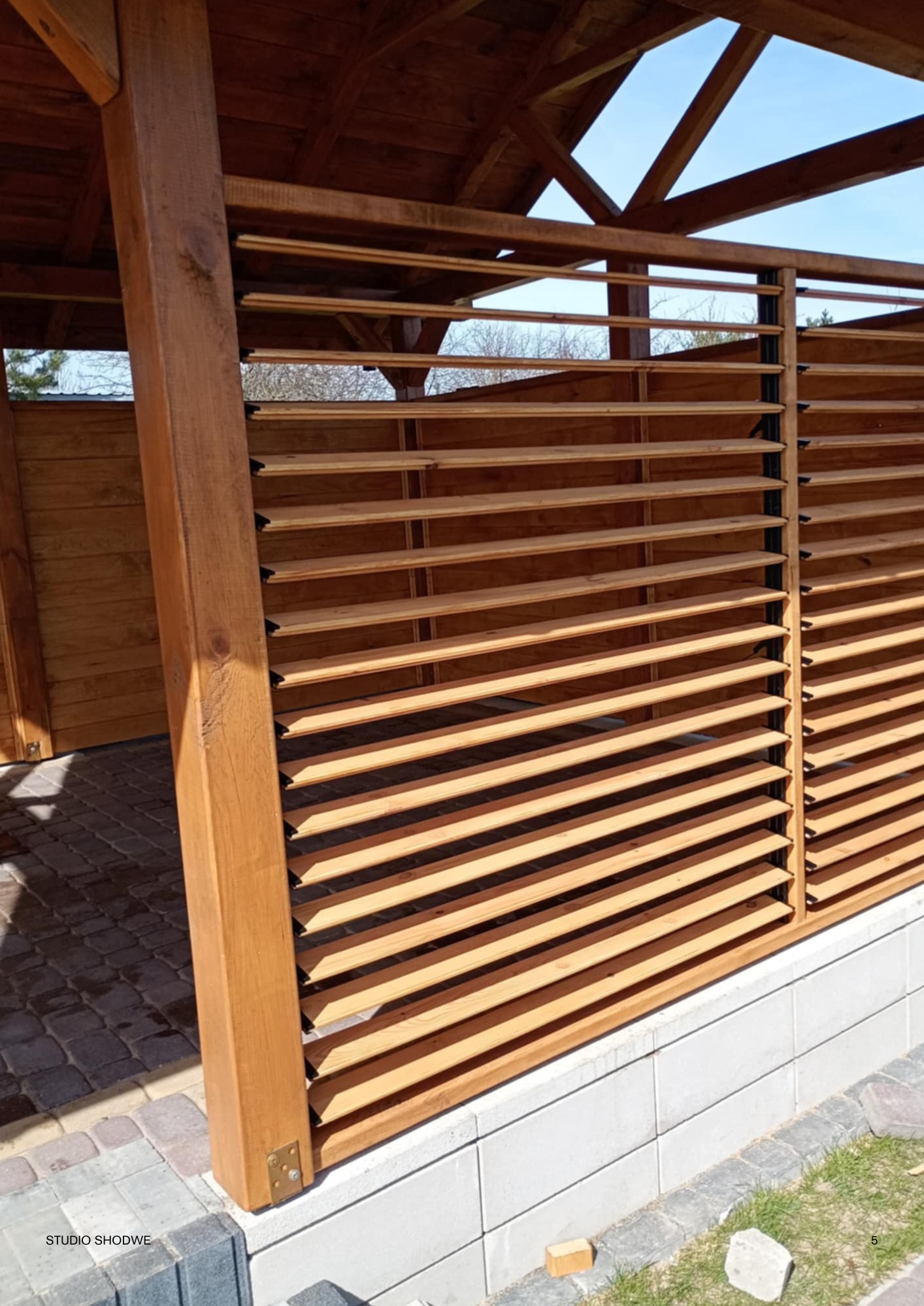 Drewniana pergola z lamelami, widok z boku, ukazujący konstrukcję i detale wykończenia. Jasne drewno, regularny układ lameli, fragment kostki brukowej w tle.
