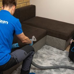 Luno Clean - Pracownik w niebieskiej koszulce z logo czyści narożnikową kanapę parownicą. Obok stoi profesjonalny sprzęt do czyszczenia tapicerki. Dywan z długim włosiem.